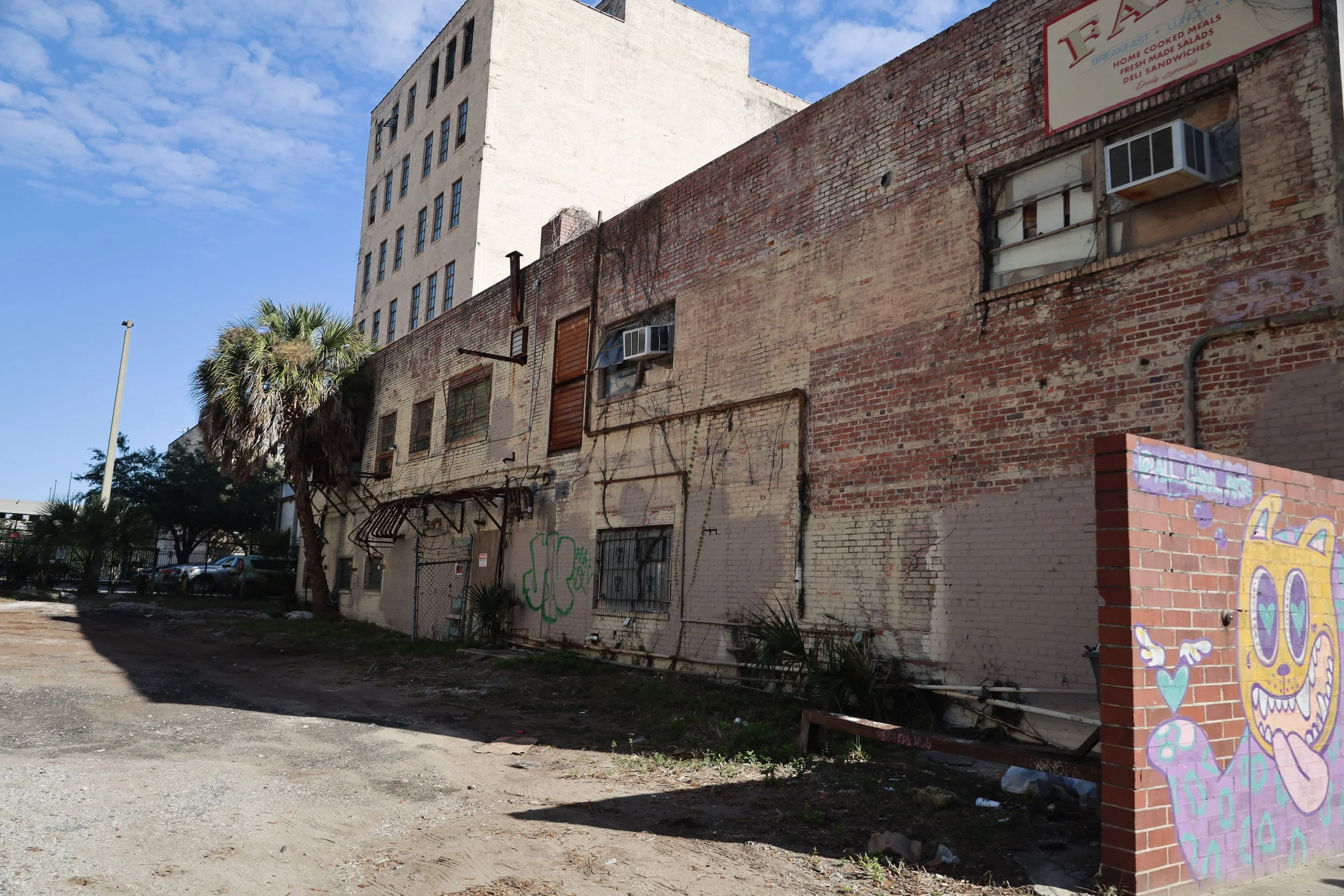 An old brick building with various air conditioning units in the windows. There is some graffiti art on a small brick wall in the foreground. A palm tree is visible on the left, and there is a taller white building in the background.