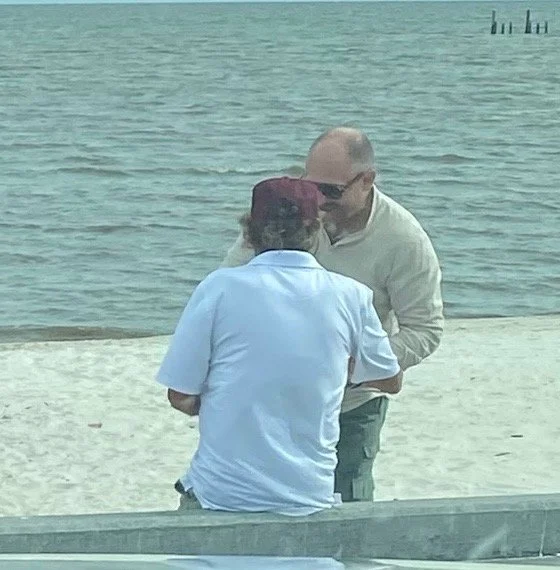 Two men face each other on a beach near the water, one wearing a white polo shirt and the other in a light-colored long sleeve shirt and sunglasses, engaging in conversation.
