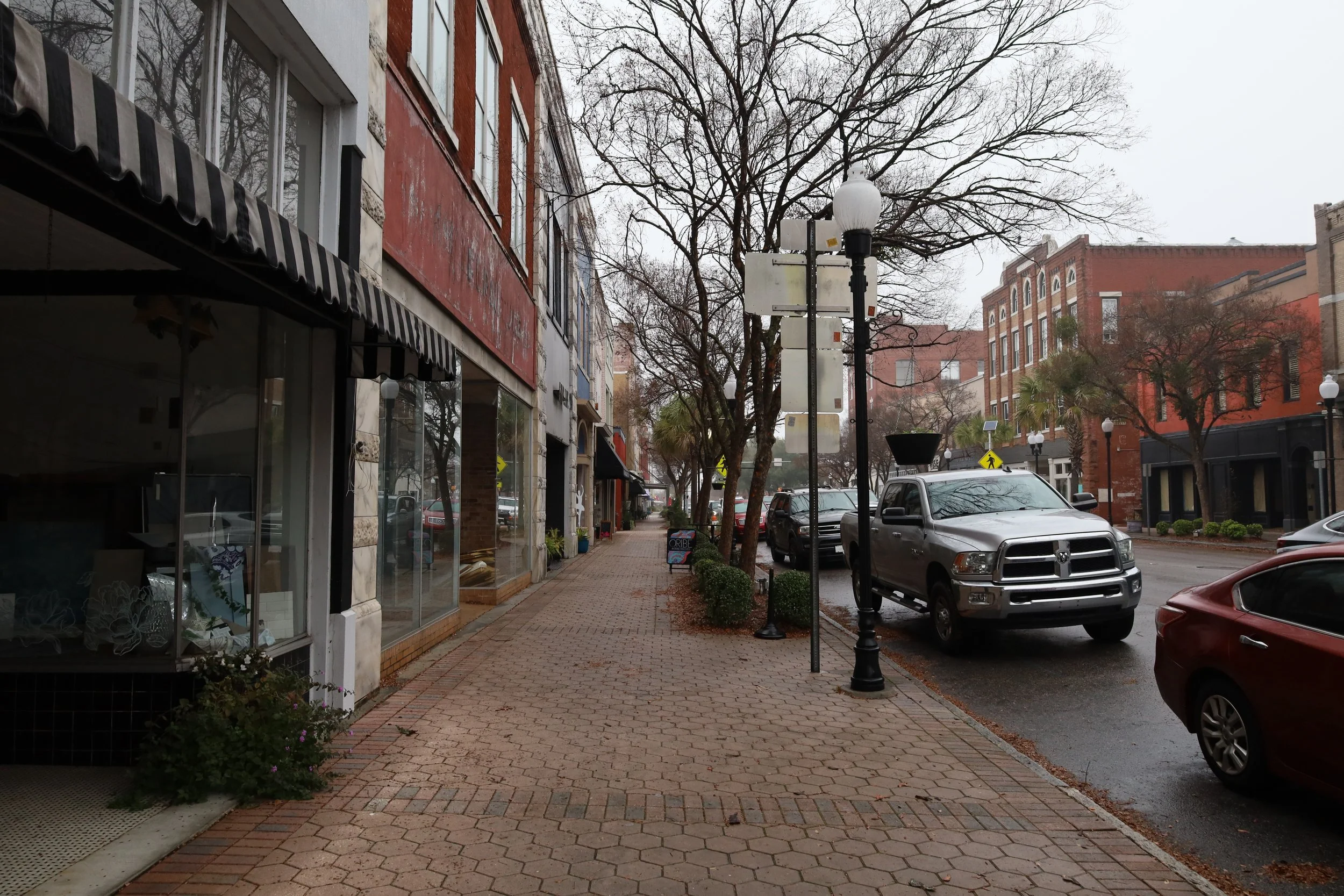 Empty sidewalk lined with storefronts on the left and parked cars on the right, with leafless trees and overcast sky in the background.