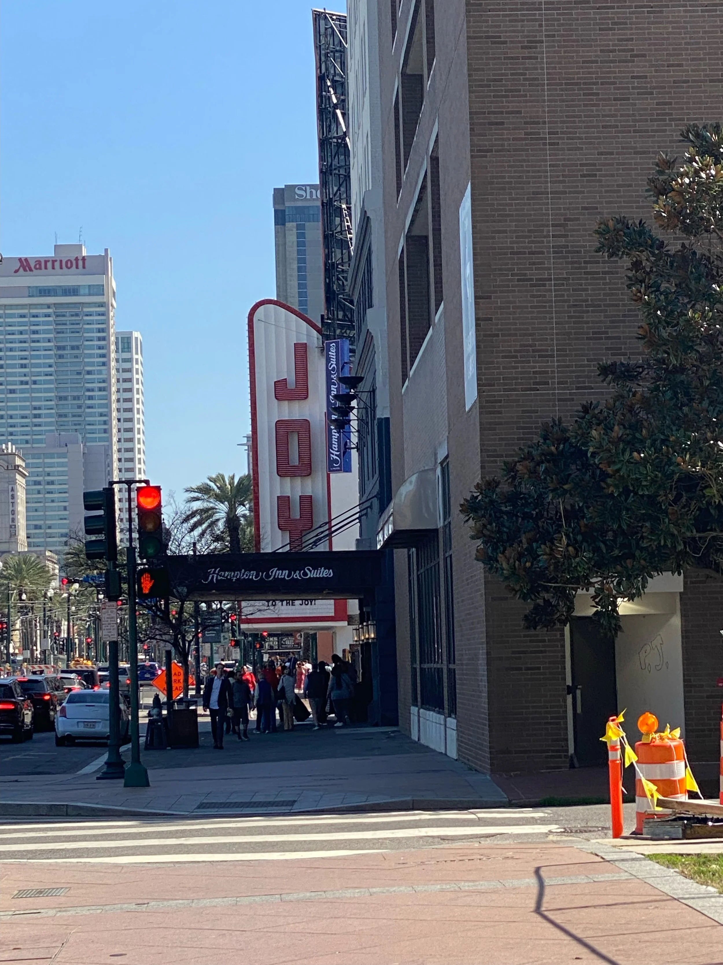 Street view in downtown with a Hilton Inn & Suites hotel entrance, people walking, traffic light, cars, tall buildings, and a construction cone. Theater is the "JOY"