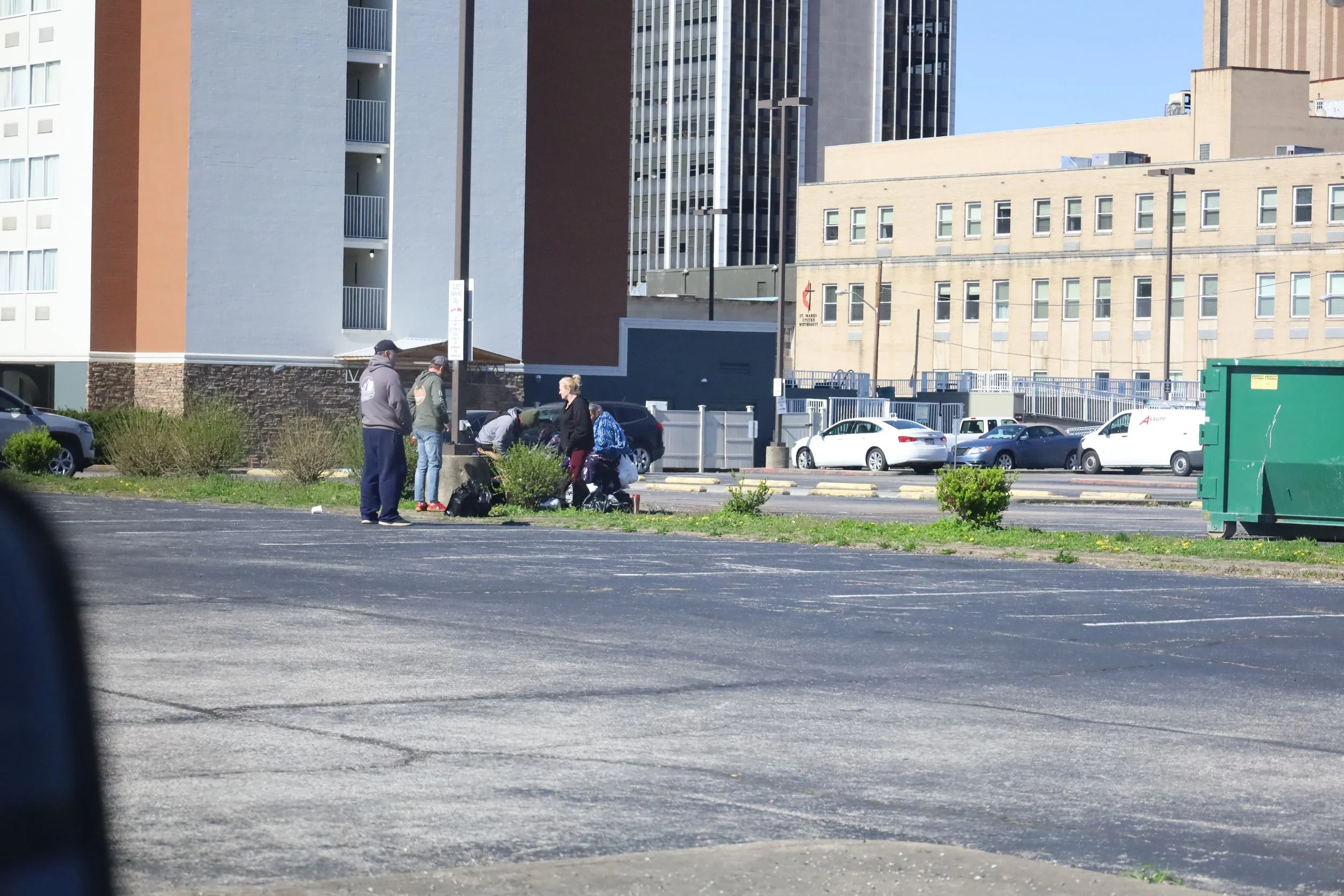 A group of people gathered around a small shrub praying in a parking lot of an urban area with tall buildings and parked cars in the background.