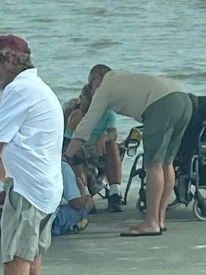 A man bending over near a wheelchair and other people at the waterfront.