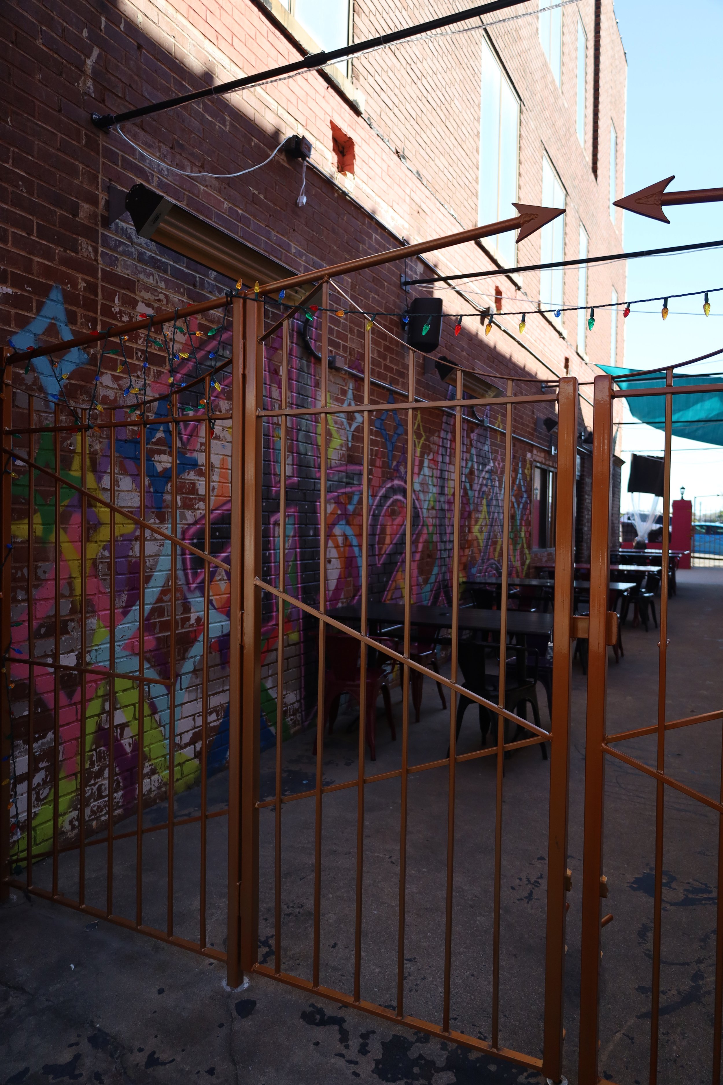 Outdoor patio area enclosed by a metal gate, with string lights hanging above, against a brick wall painted with colorful graffiti art, and with tables and chairs arranged for seating.