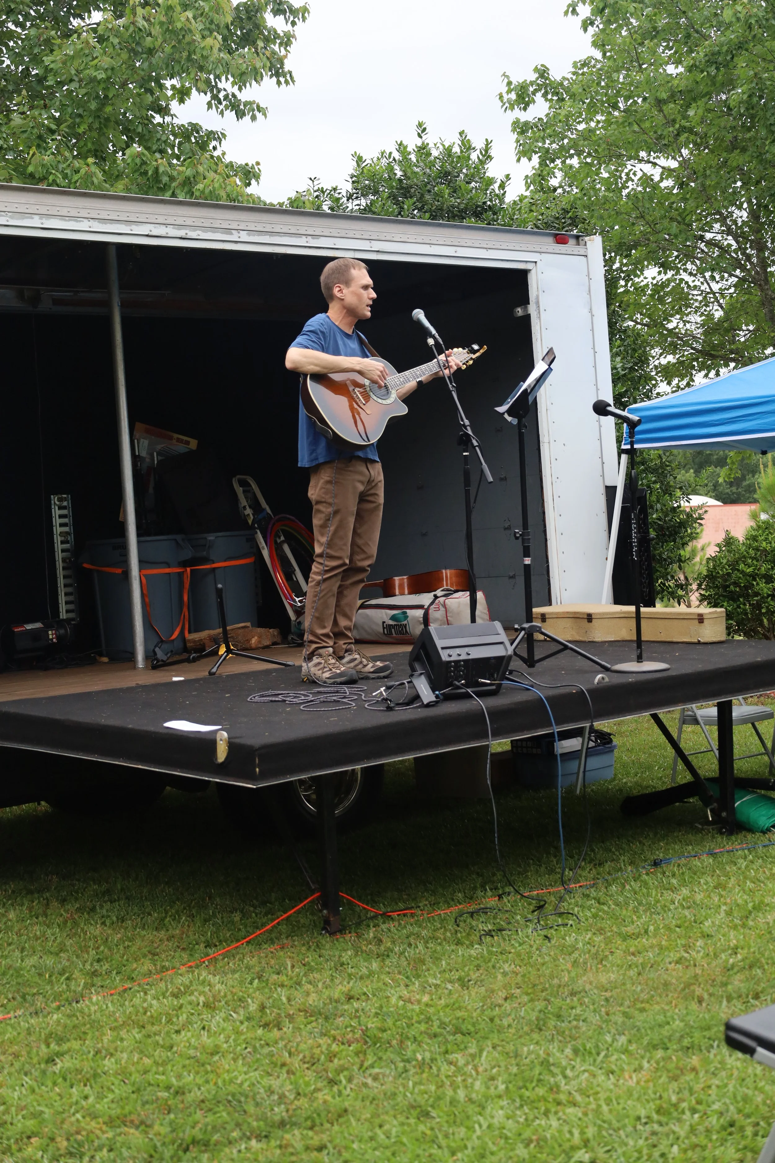 A man playing an acoustic guitar on a small outdoor stage with trees in the background leads a large group of people in worship.