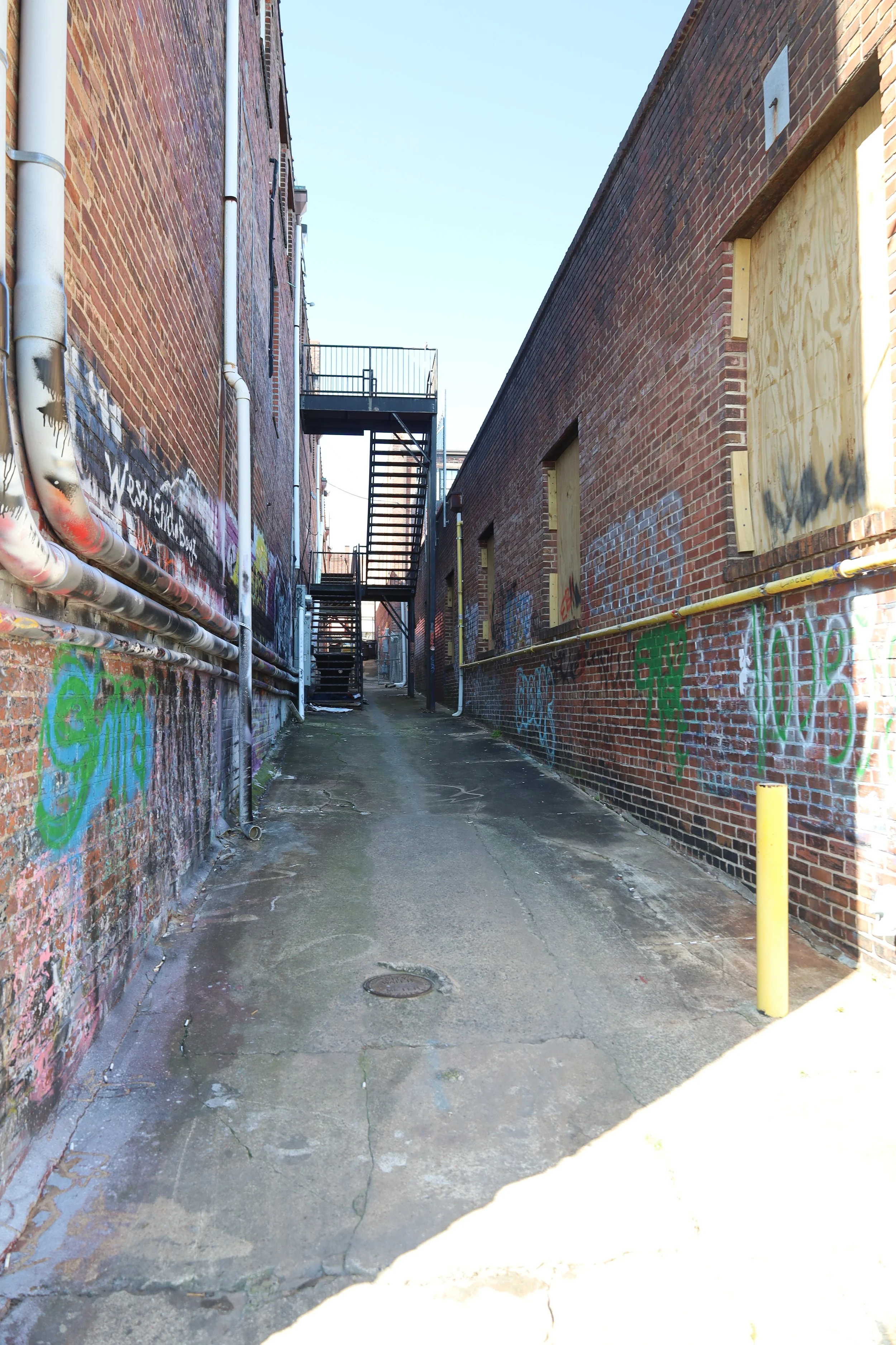 An alleyway between two brick buildings with graffiti on the walls, metal pipes, a yellow bollard, and an outdoor metal staircase leading to a fire escape.