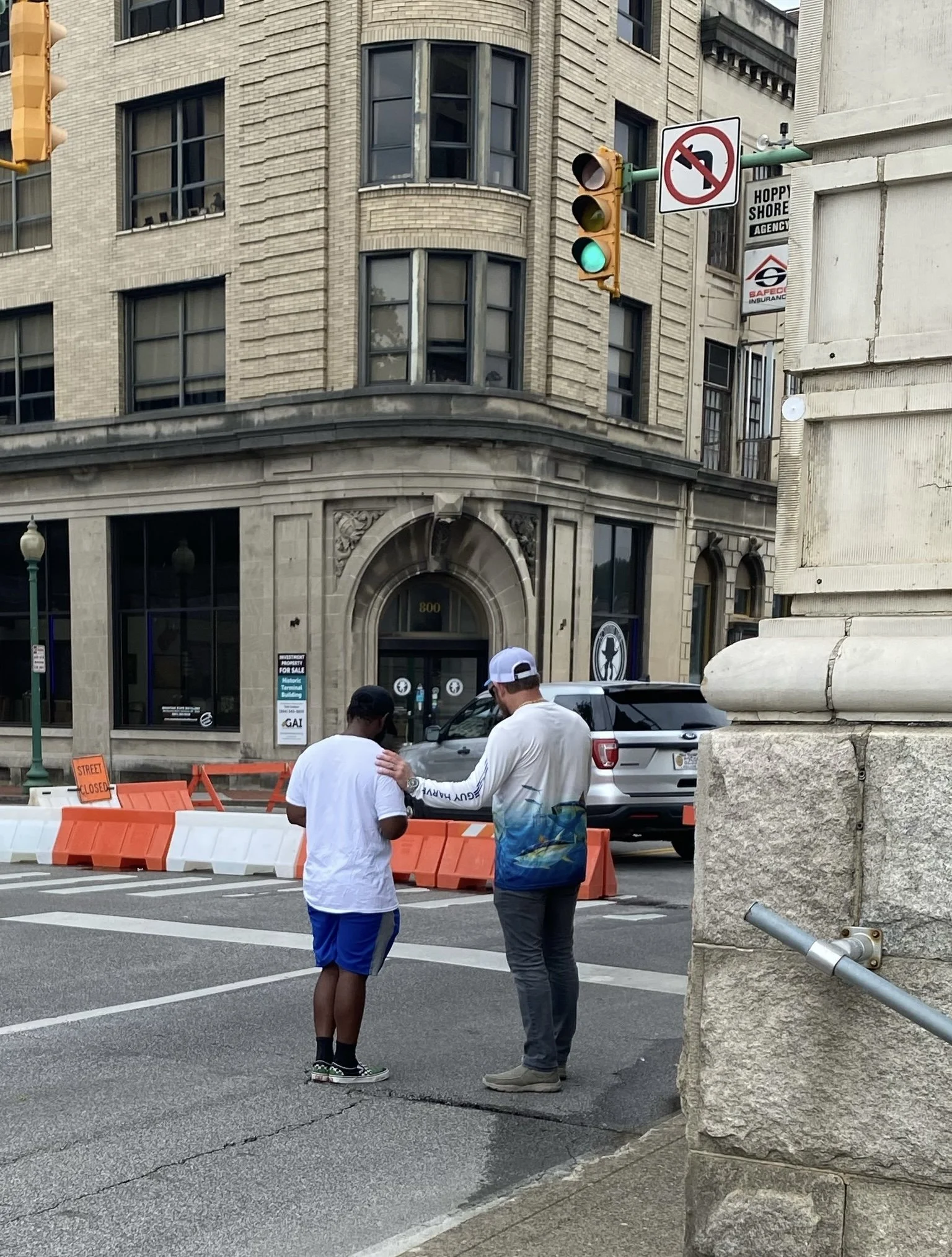 Two men standing on the street conversing, with one wearing a white shirt and black cap, and the other wearing a white long-sleeve shirt with a colorful design, gray pants, and a white cap. Behind them are orange and white construction barriers, a gr