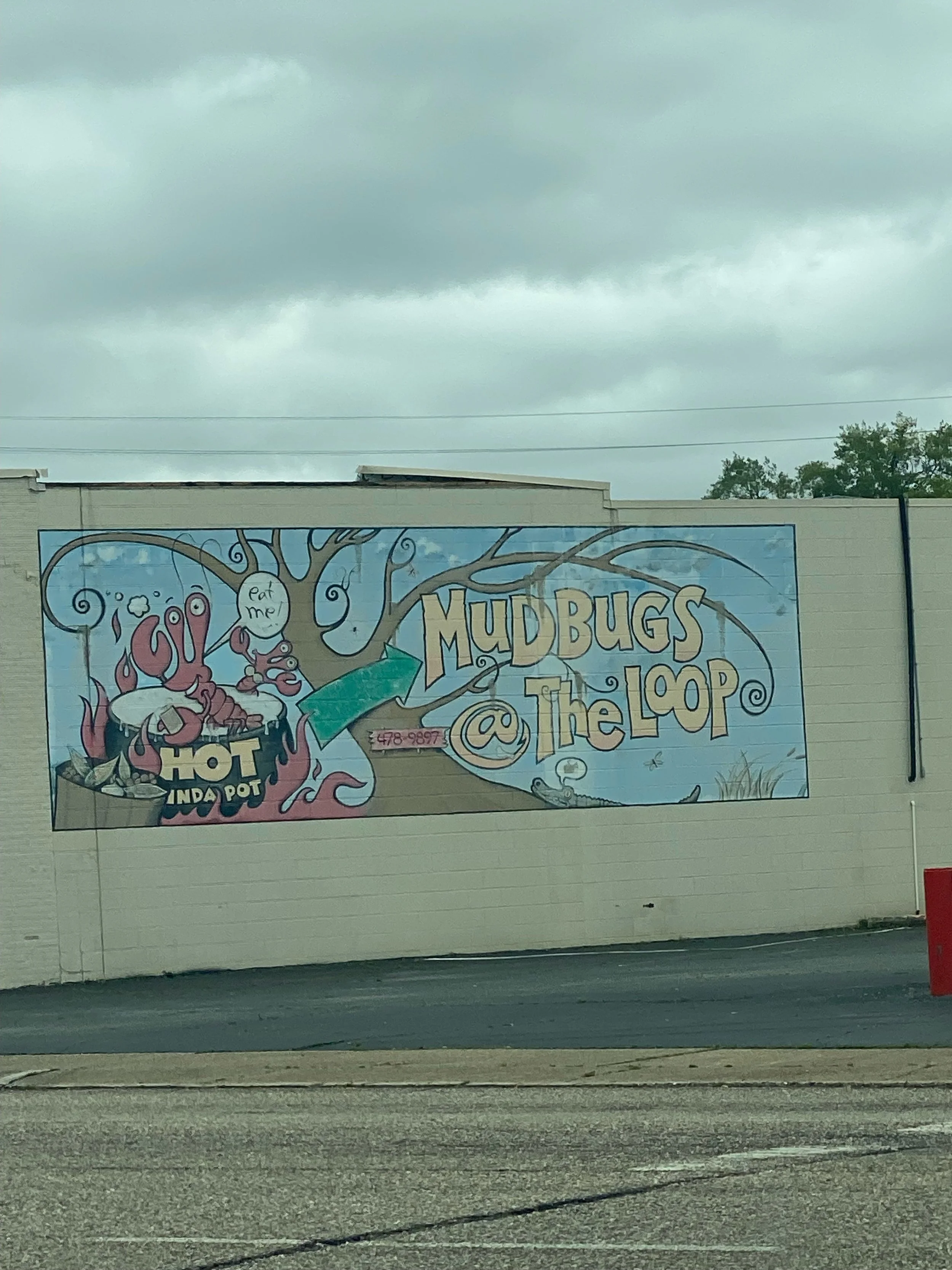A mural on a building wall advertising Mud Bugs @ The Loop, featuring a cartoon crawfish with a speech bubble saying "Eat me!" next to a steaming bowl labeled "Hot IND A POT" and a cartoon mouse, with cloudy sky above.