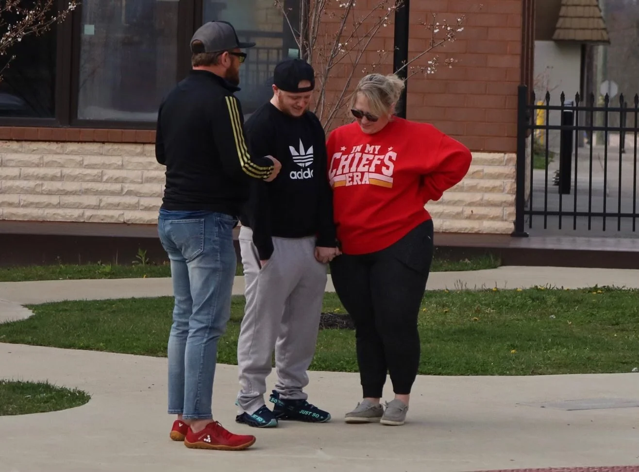 Three people standing outdoors on a sidewalk, smiling and looking down praying. Two men and one woman, casual clothing, with a building and a small tree in the background.