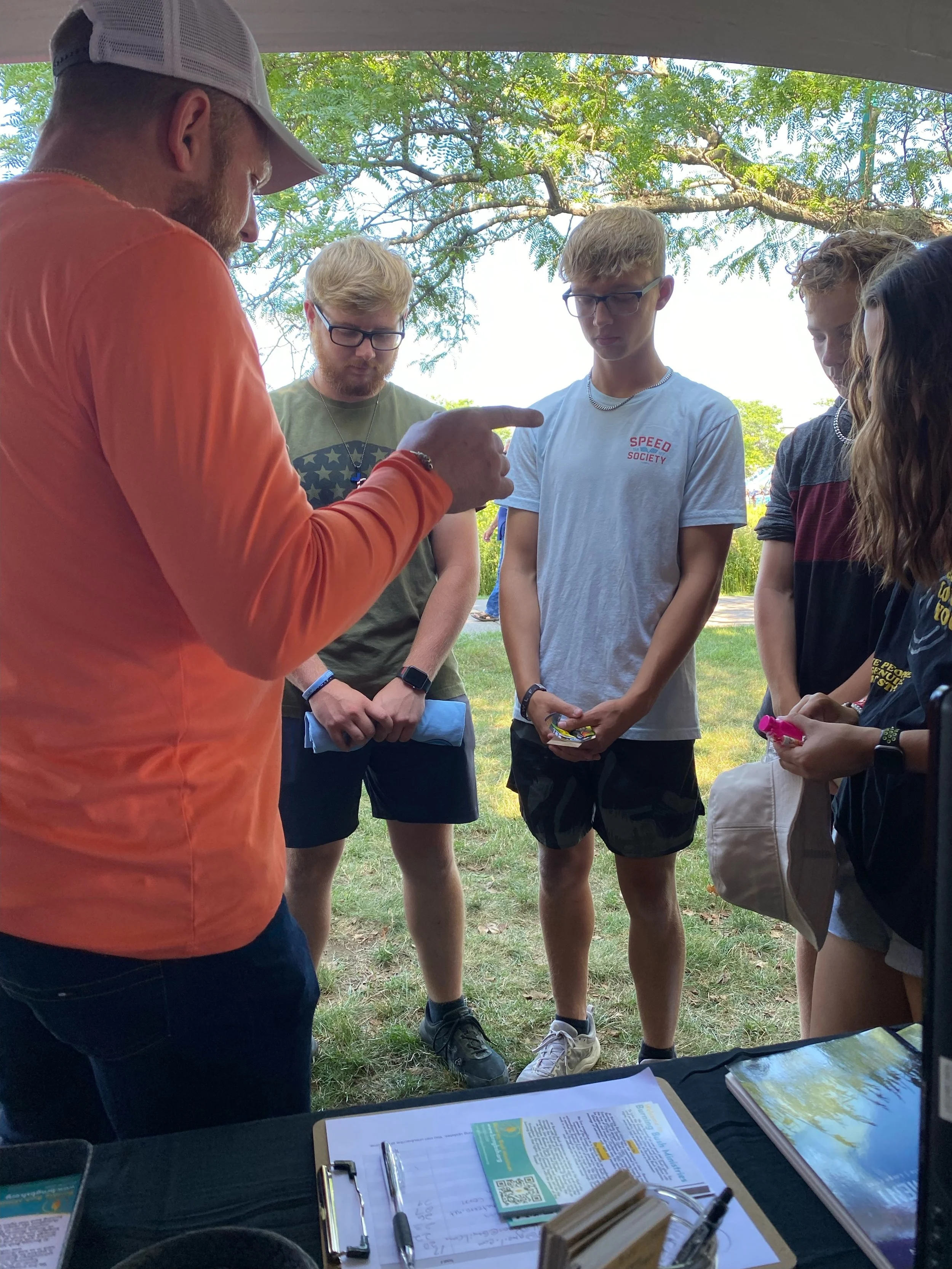 An man with a beige cap and orange shirt talking to a group of five teenagers standing outdoors under a tree.