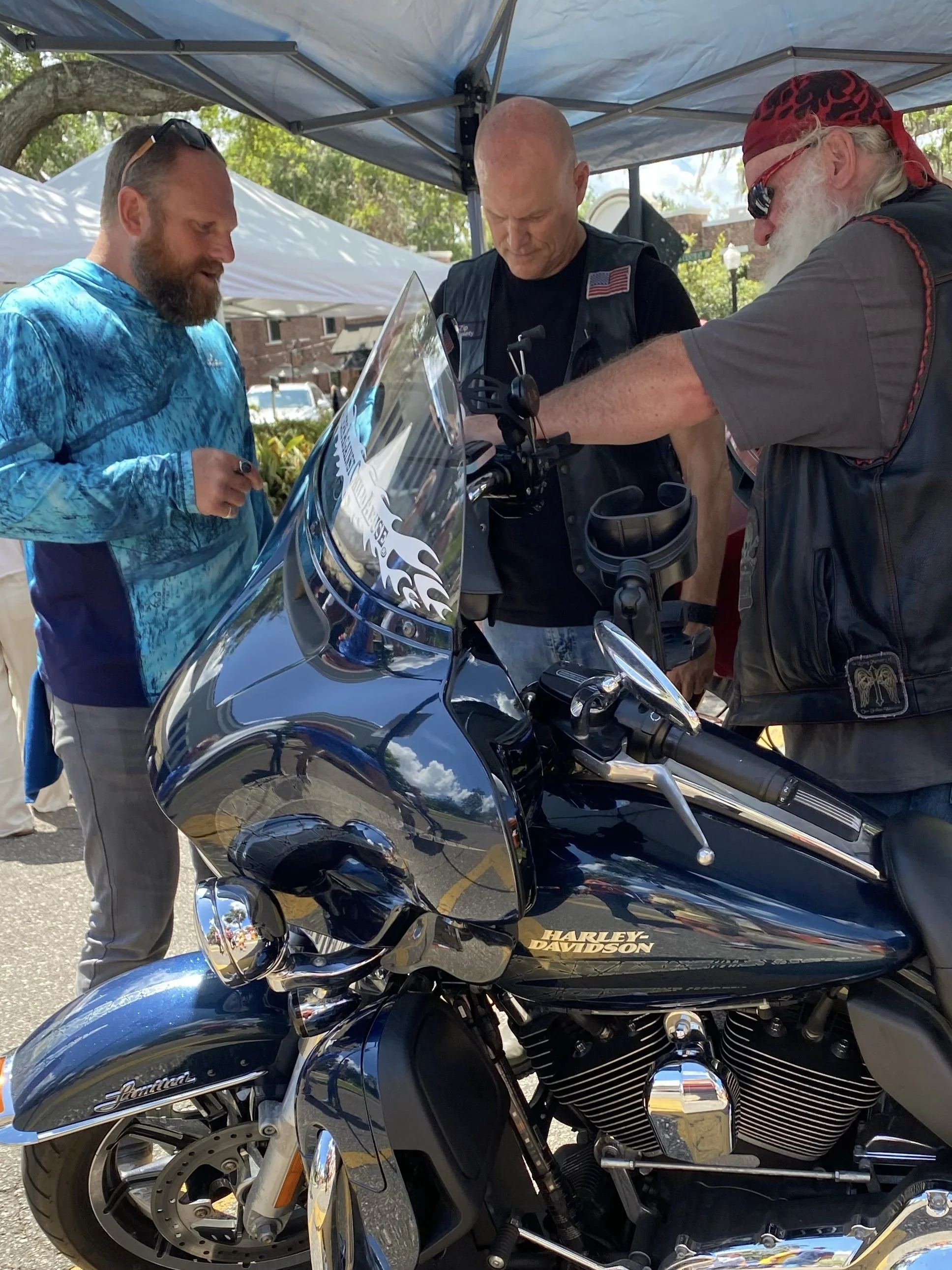 Three men gathered around a black Harley Davidson motorcycle under a canopy at an outdoor event. The men are praying.