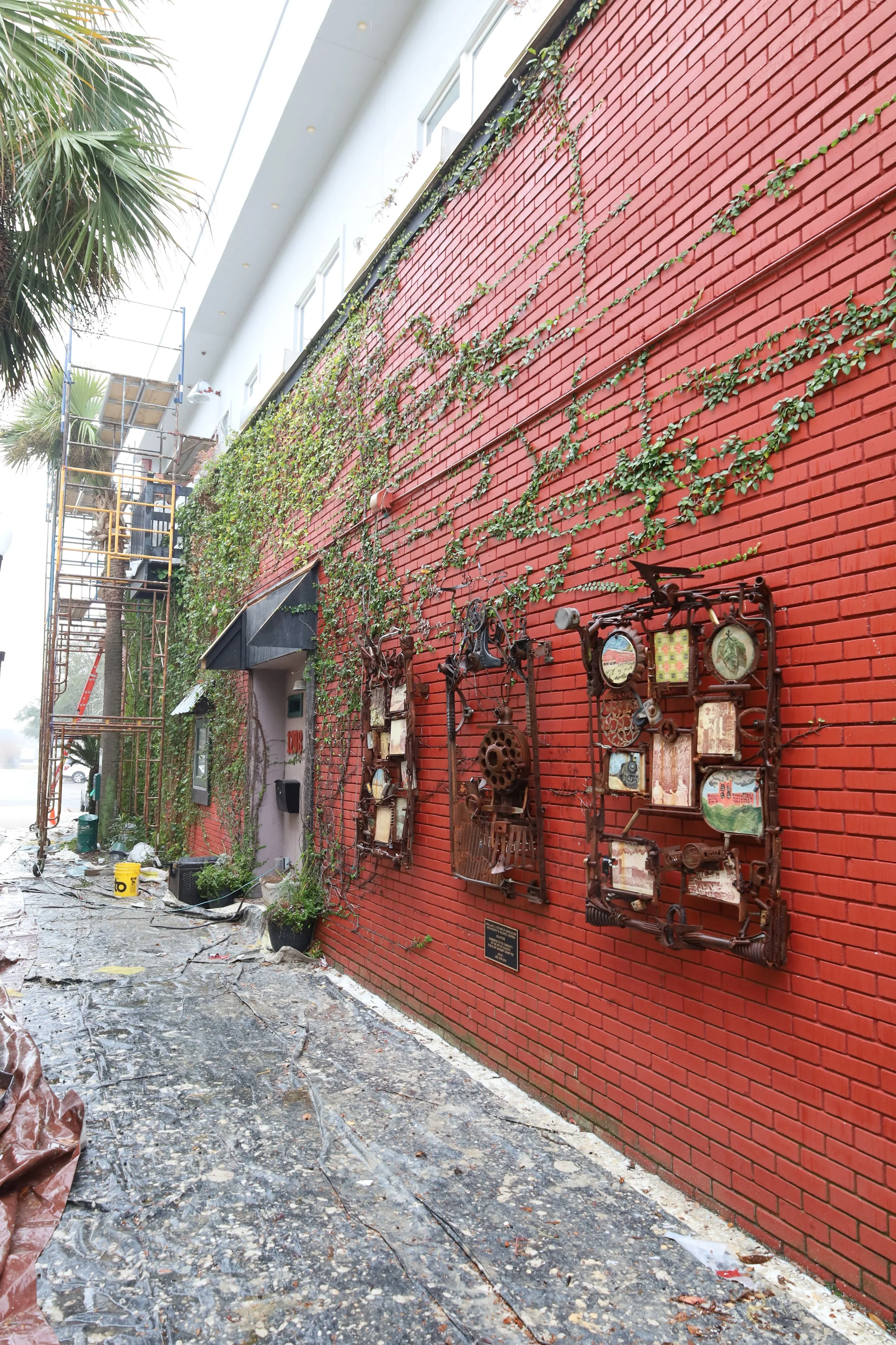 Decorative wall art with metal frames, gears, and clocks on a red brick wall, partially covered by a vine, in an outdoor urban space under construction with scaffolding, scattered tools, and supplies on the ground.
