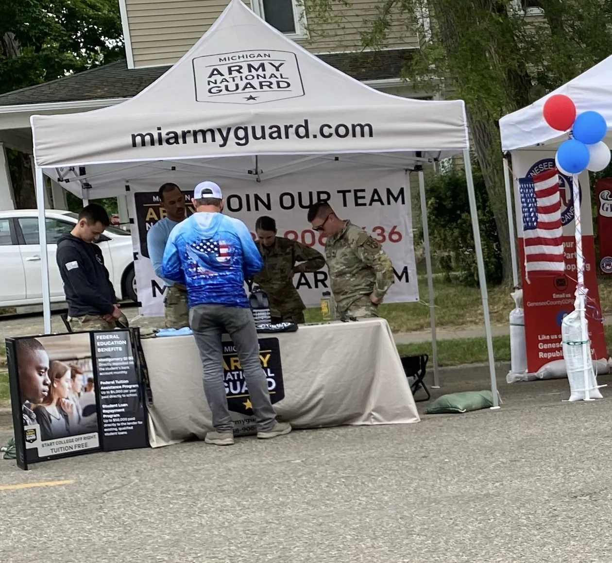A booth under a white canopy with the Michigan Army National Guard logo and website. Several military personnel and a civilian are interacting at the booth, which features informational posters and a banner inviting people to join the team. A small A