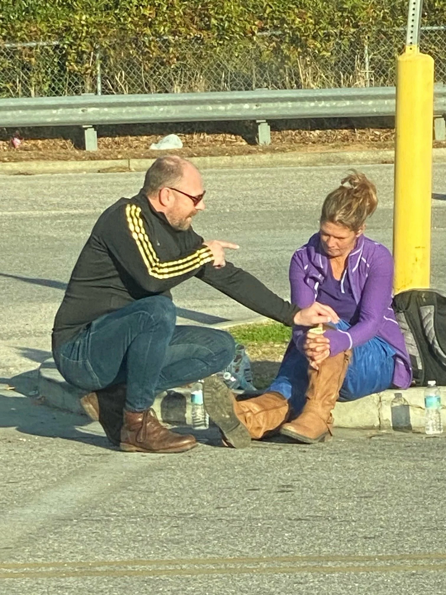A man is praying with a woman who is sitting on the sidewalk. They are near a street corner with a yellow pole and a guardrail in the background.