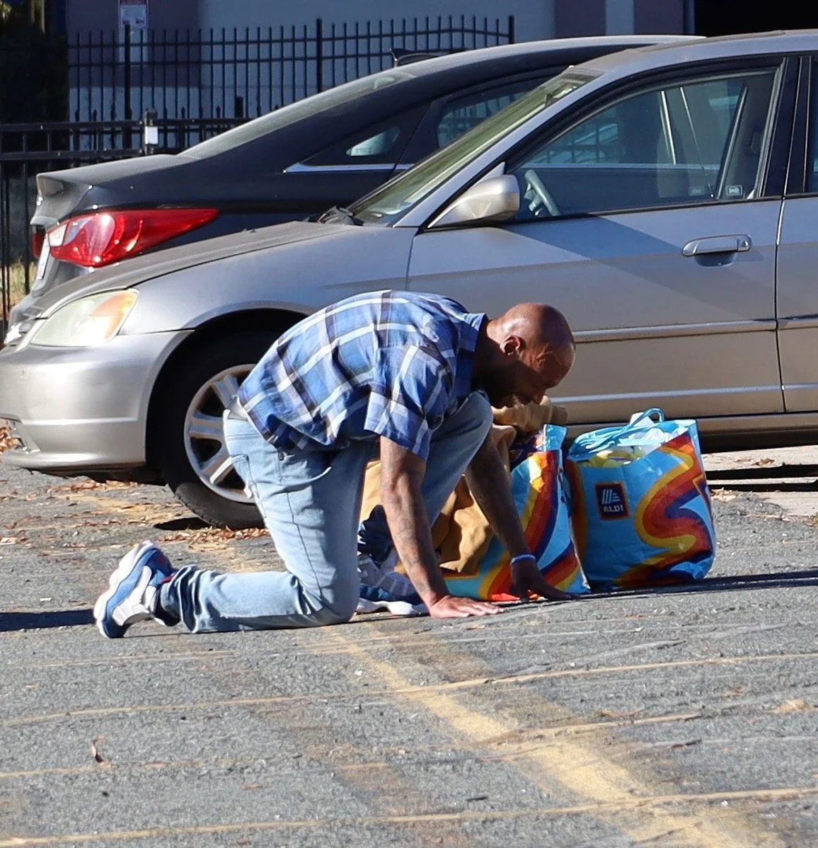 A man kneeling on the pavement with his hands on the ground, next to colorful shopping bags, in front of parked cars in a parking lot.