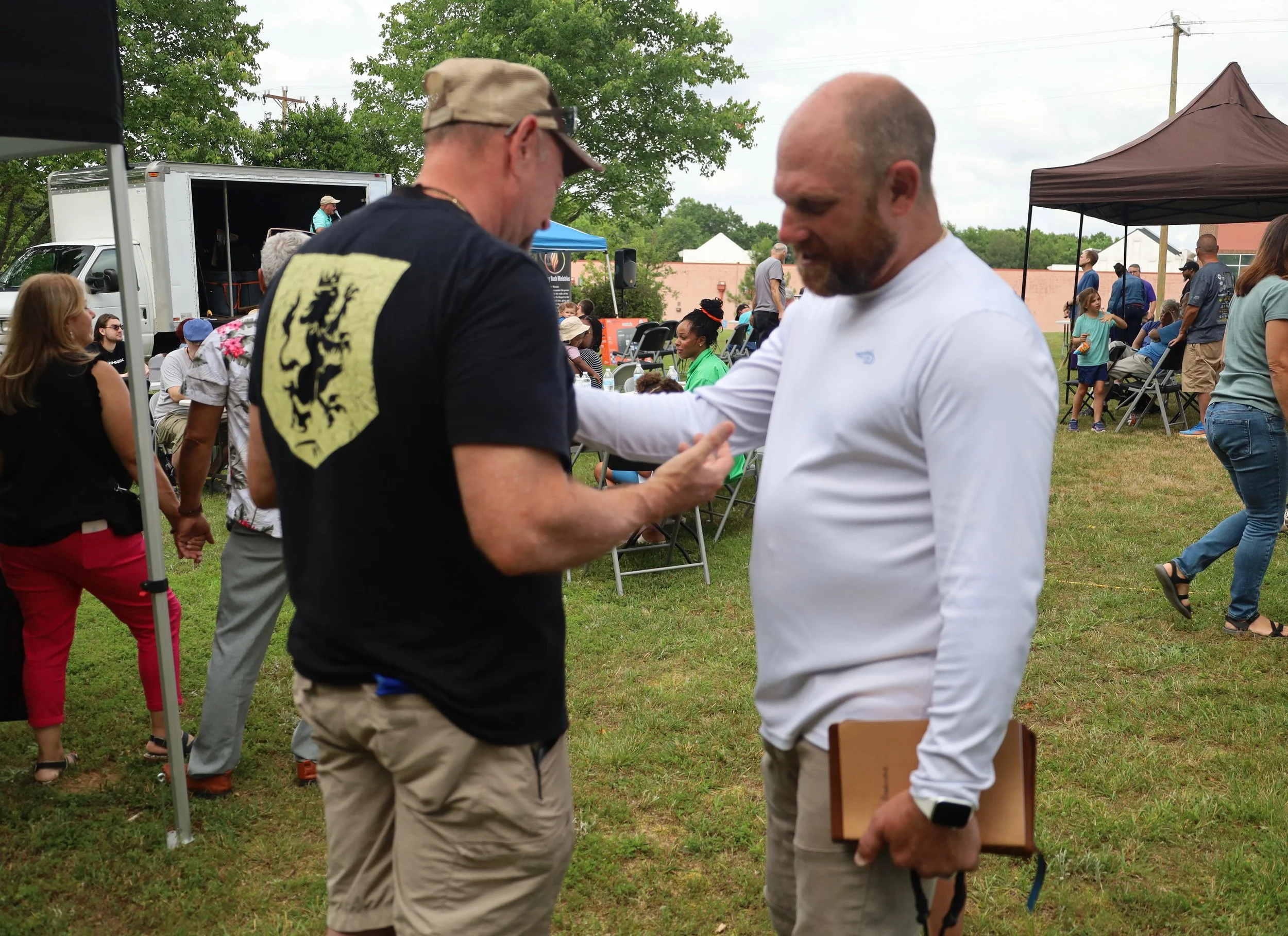 Two men shaking hands at an outdoor gathering, one holding a notebook, with several people sitting and standing under tents and in the background.