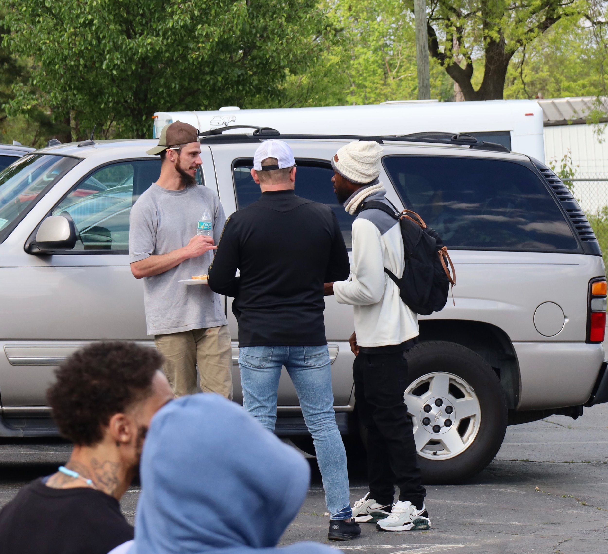 Three men, one in a gray t-shirt and beige pants, another in a black shirt and jeans, and the third in a white jacket with black pants, stand near a silver SUV, talking and sharing food, with a group of people sitting in the foreground in an outdoor 