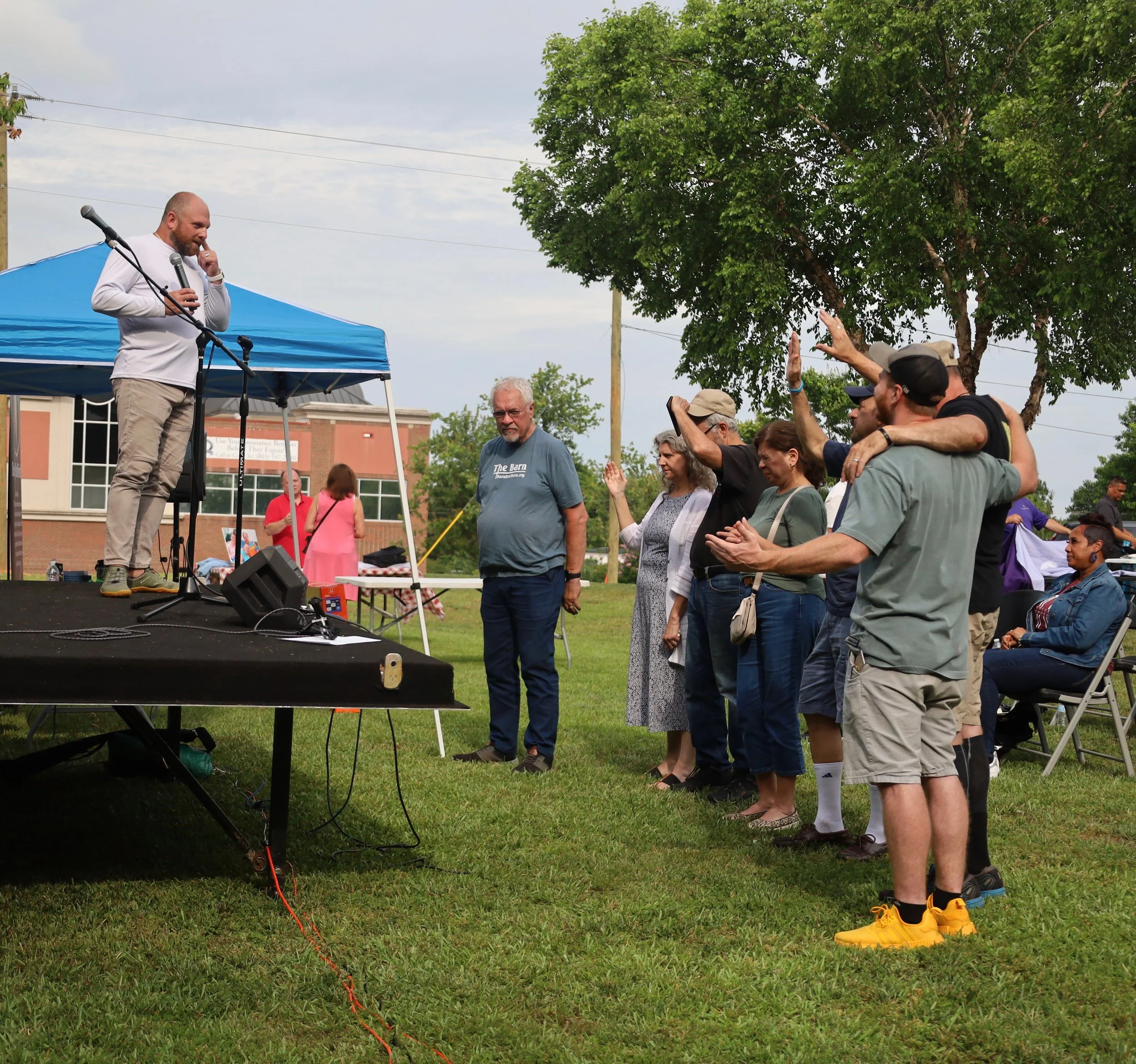 A man speaking on a stage outdoors at a community event, with a group of people standing in front of him, some raising their hands. The setting includes a blue canopy tent and a grassy area with trees and a brick building in the background.