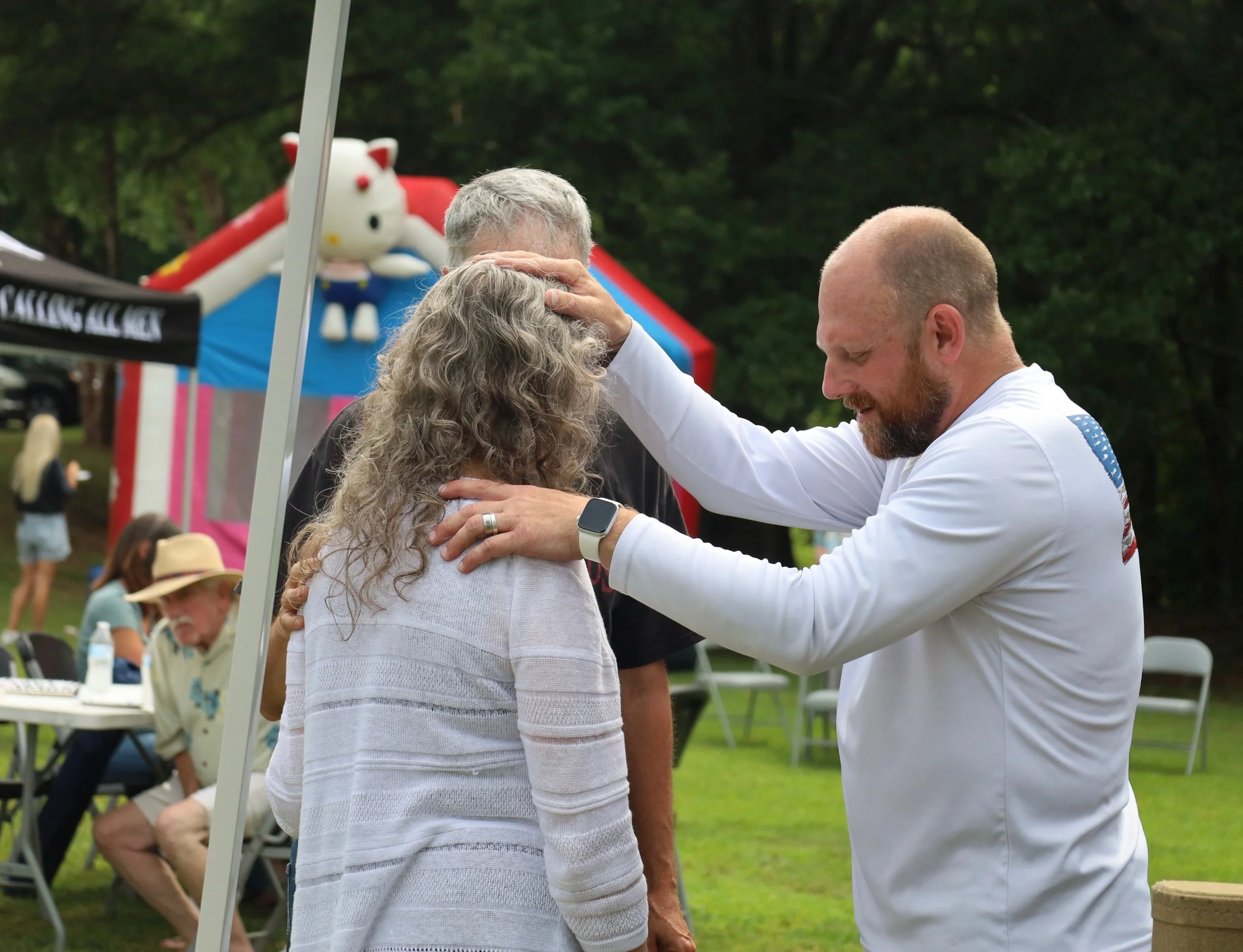 A man is praying over a woman with curly gray hair at an outdoor event, with a bouncy castle in the background.