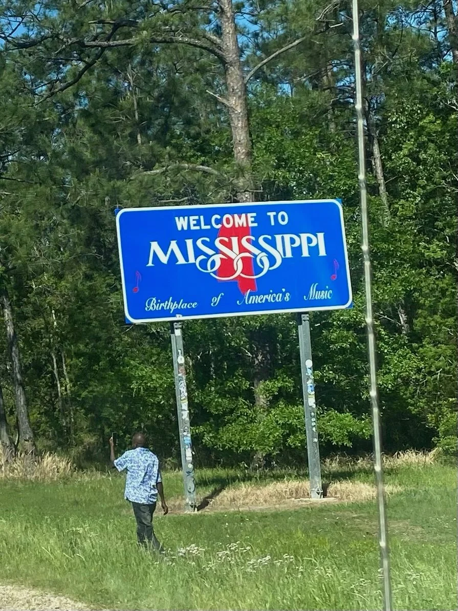 A man walking away from the camera on a grassy area, towards a large blue sign that says "Welcome to Mississippi" with a red and white state outline and the words "Birthplace of America's Music". There are trees in the background.