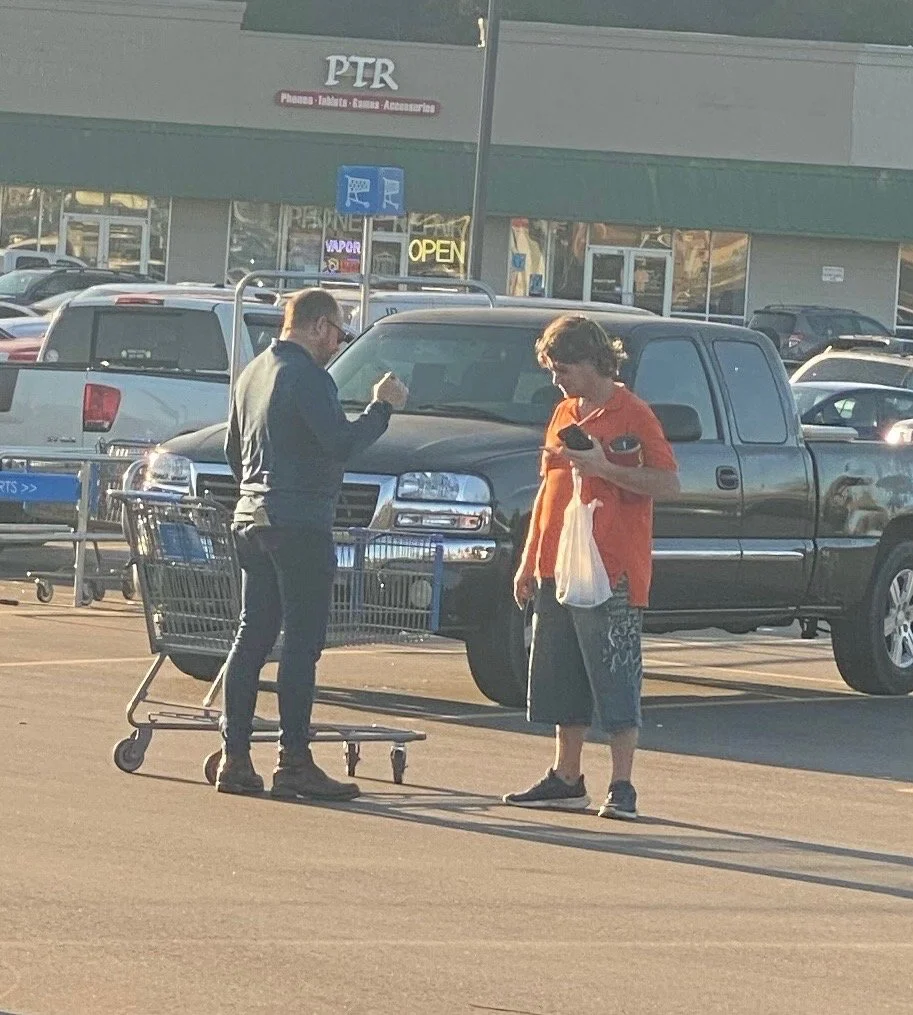 Two men standing in a parking lot, one with a shopping cart, engaged in conversation. The man on the left is wearing sunglasses, a leather jacket, and jeans, while the man on the right is holding a plastic bag and wearing a red shirt and shorts. Seve