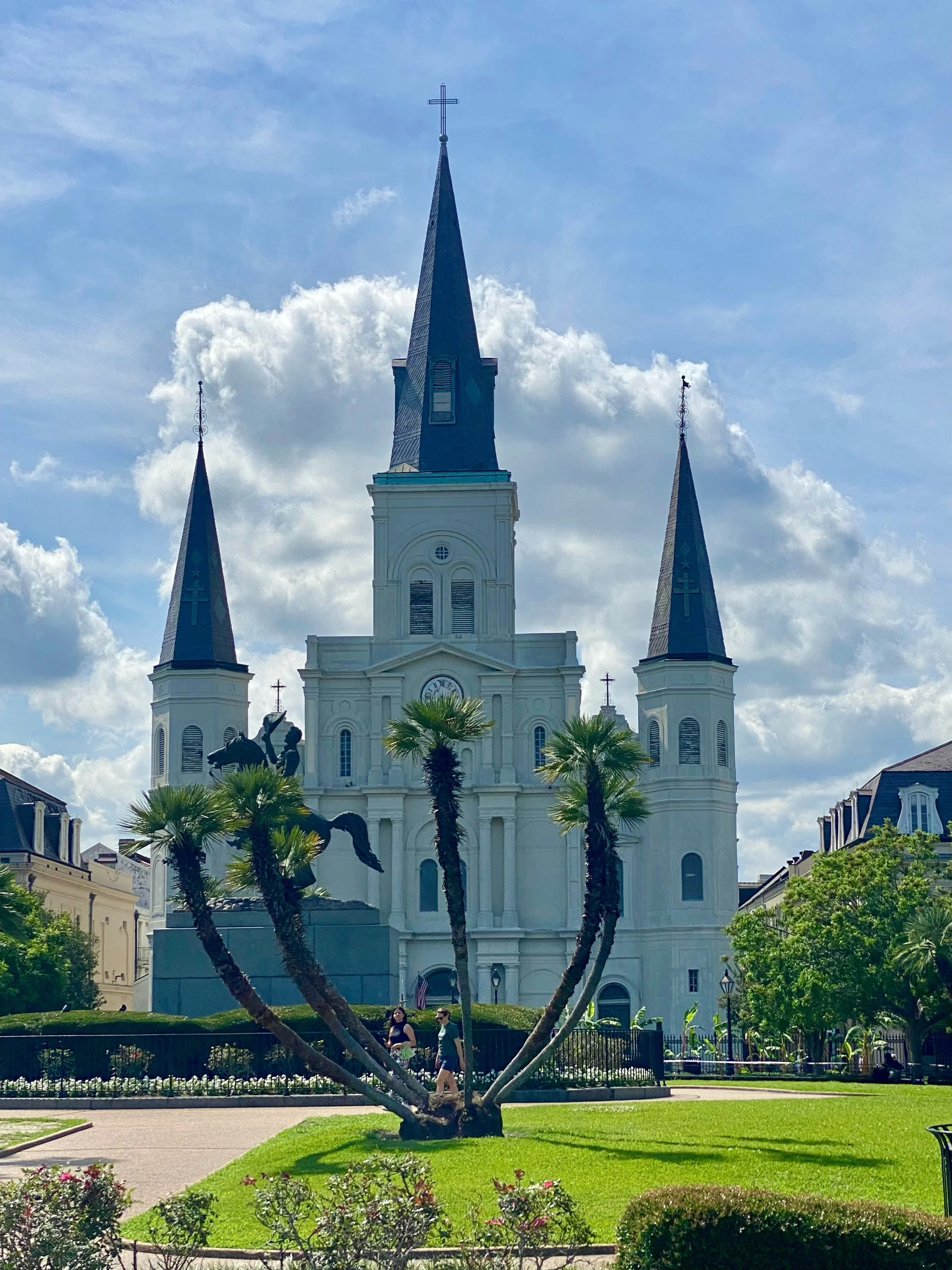 A large white cathedral with three tall spires, set in a park with palm trees and a statue of a person on a horse in front.