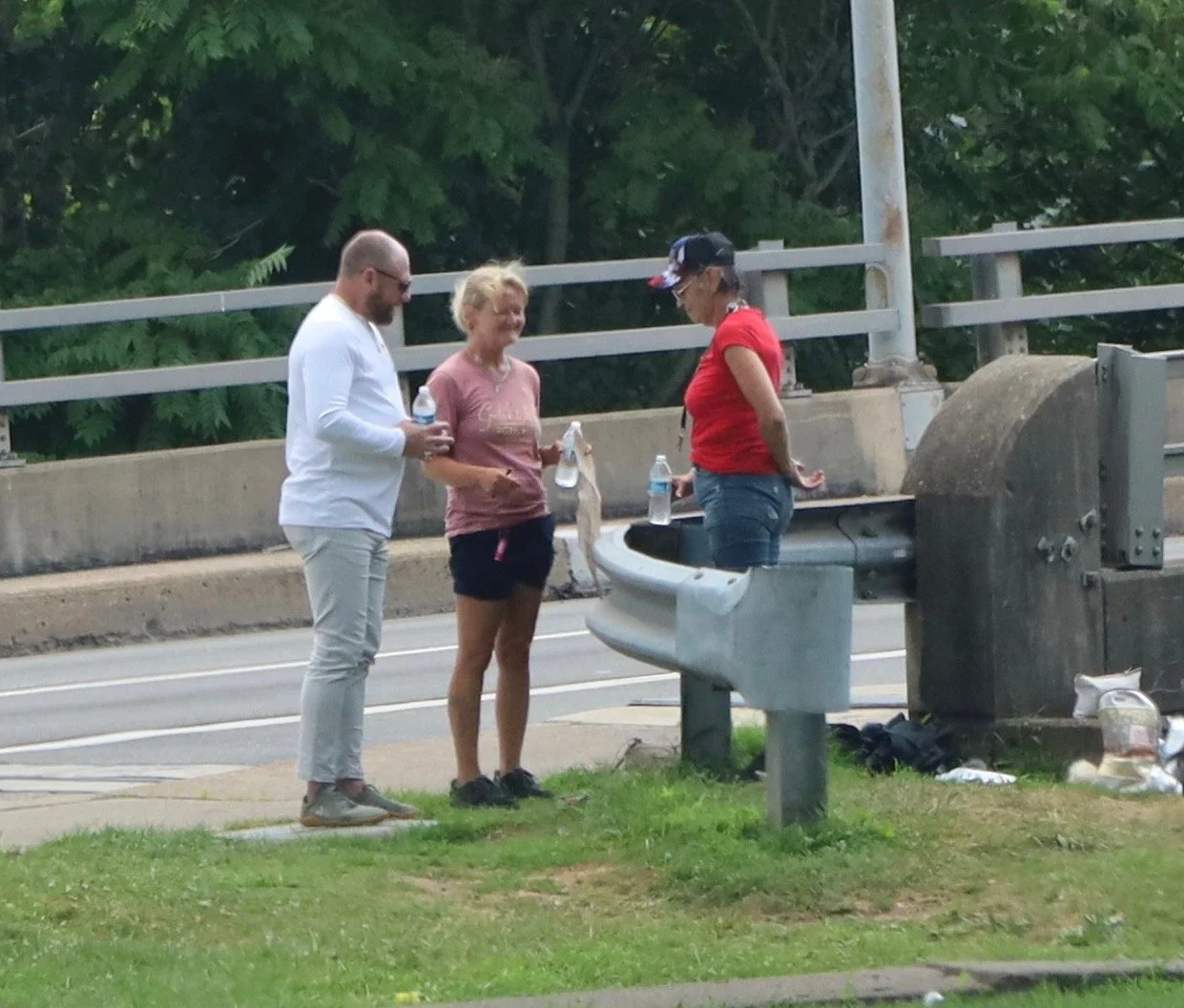 Three people standing by a concrete barrier are given water, on a roadside next to a grassy area with trees.