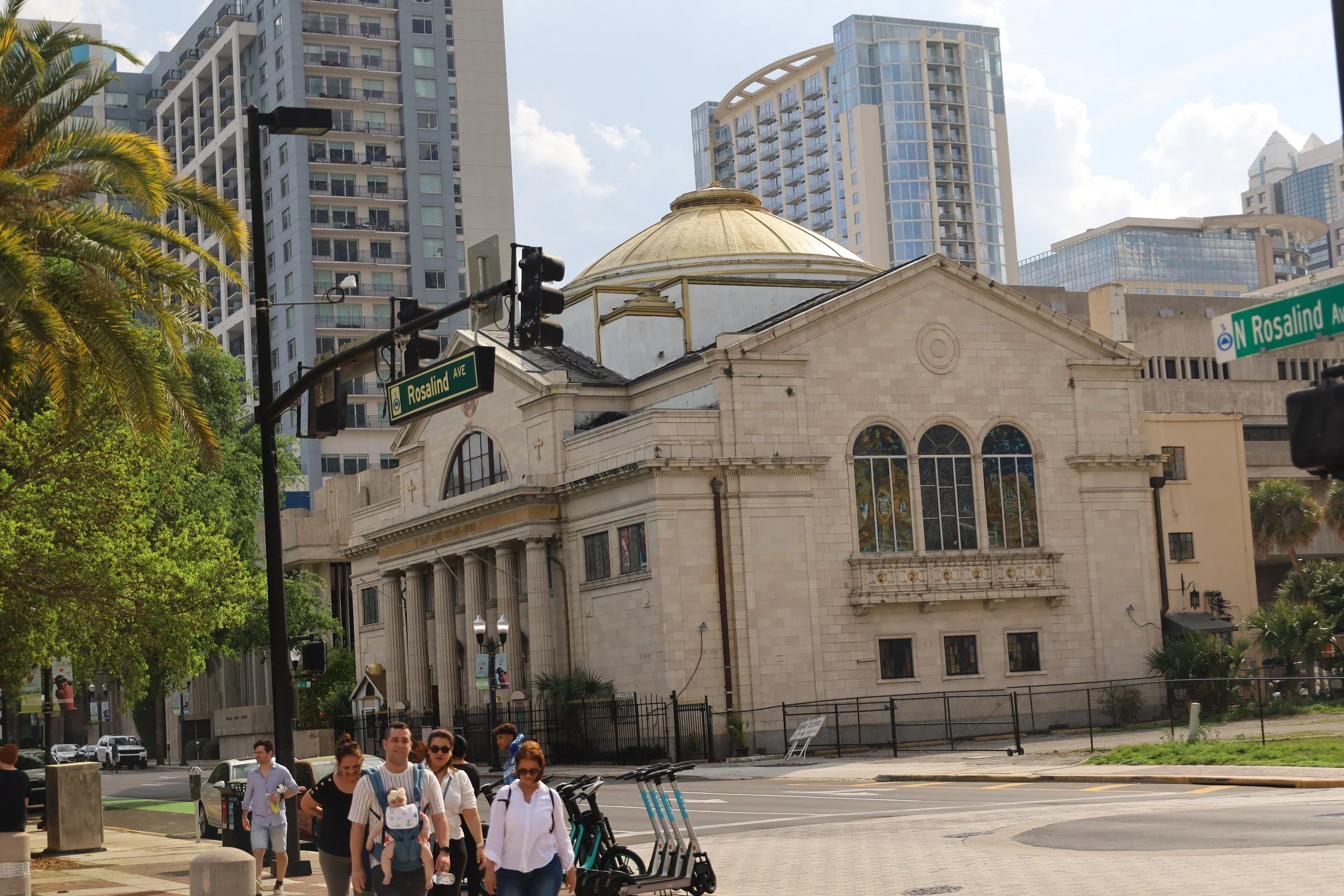 A group of people walking on a city sidewalk near a historic church with stained glass windows. Modern skyscrapers are in the background, and street signs indicate the intersection.