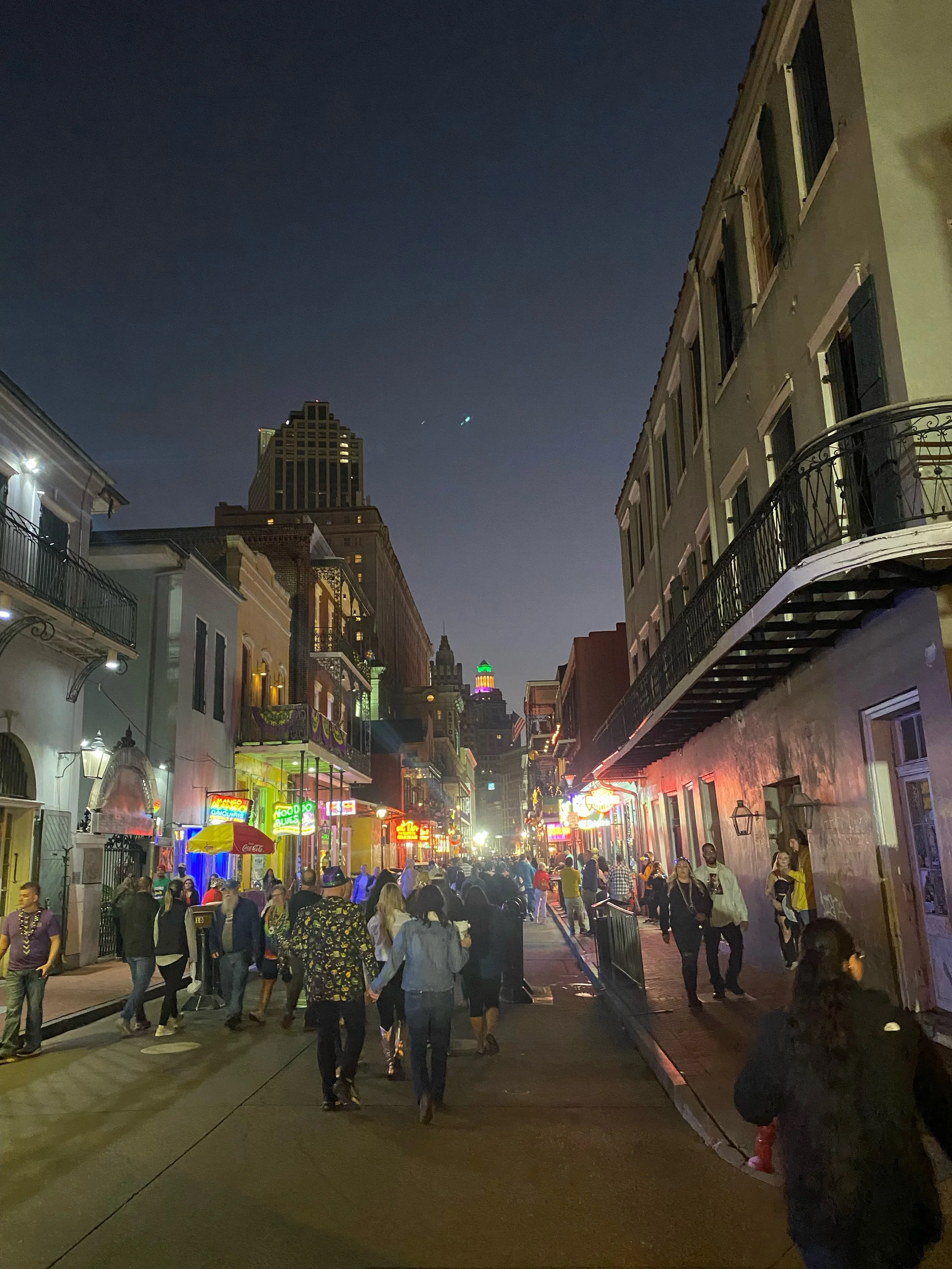 Nighttime street scene in New Orleans with neon signs, crowded sidewalk, and historic buildings.