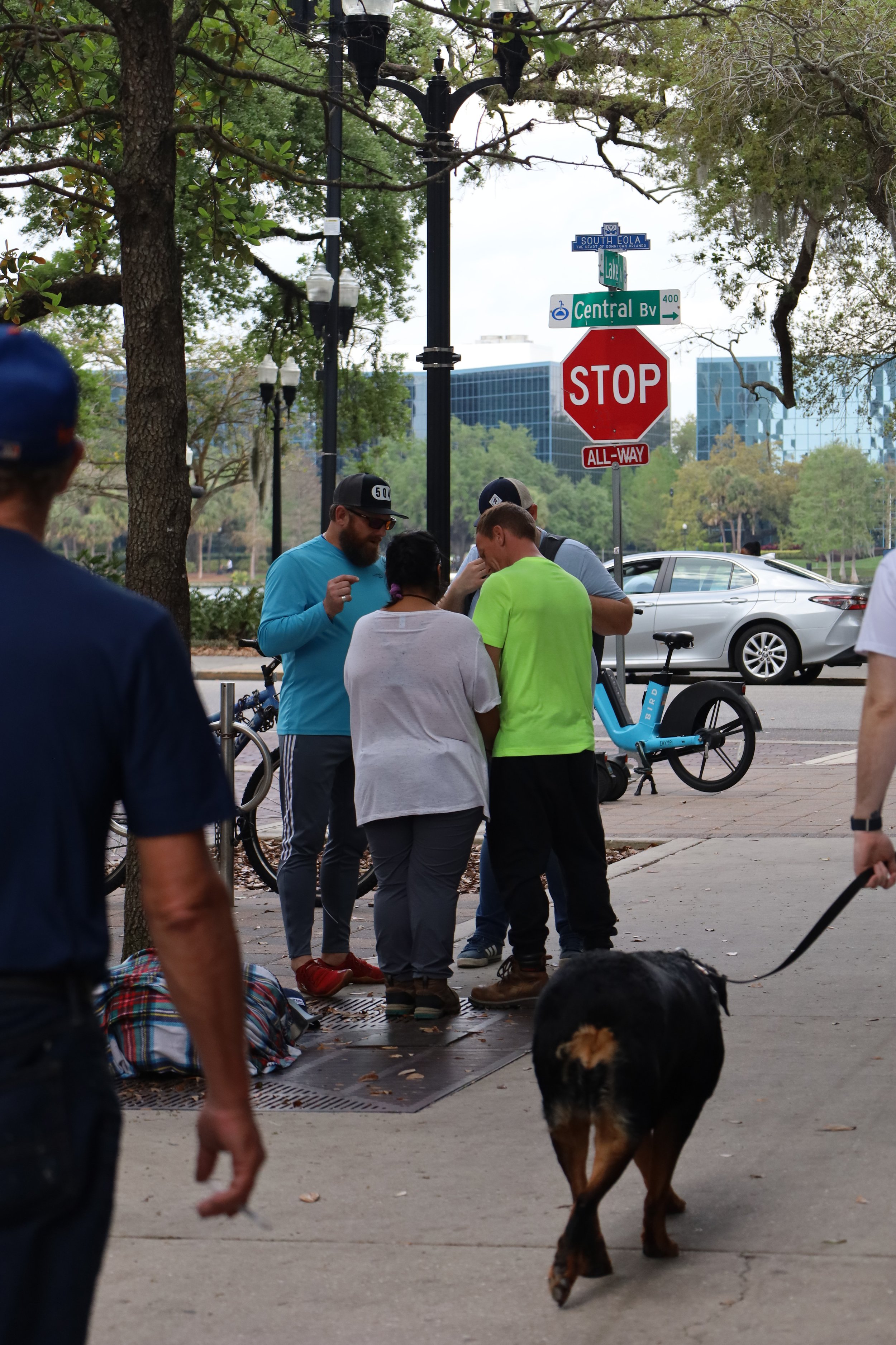 Group of people on a city sidewalk gathered around praying, with bicycles, a parked dog on a leash, trees, and street signs in the background.