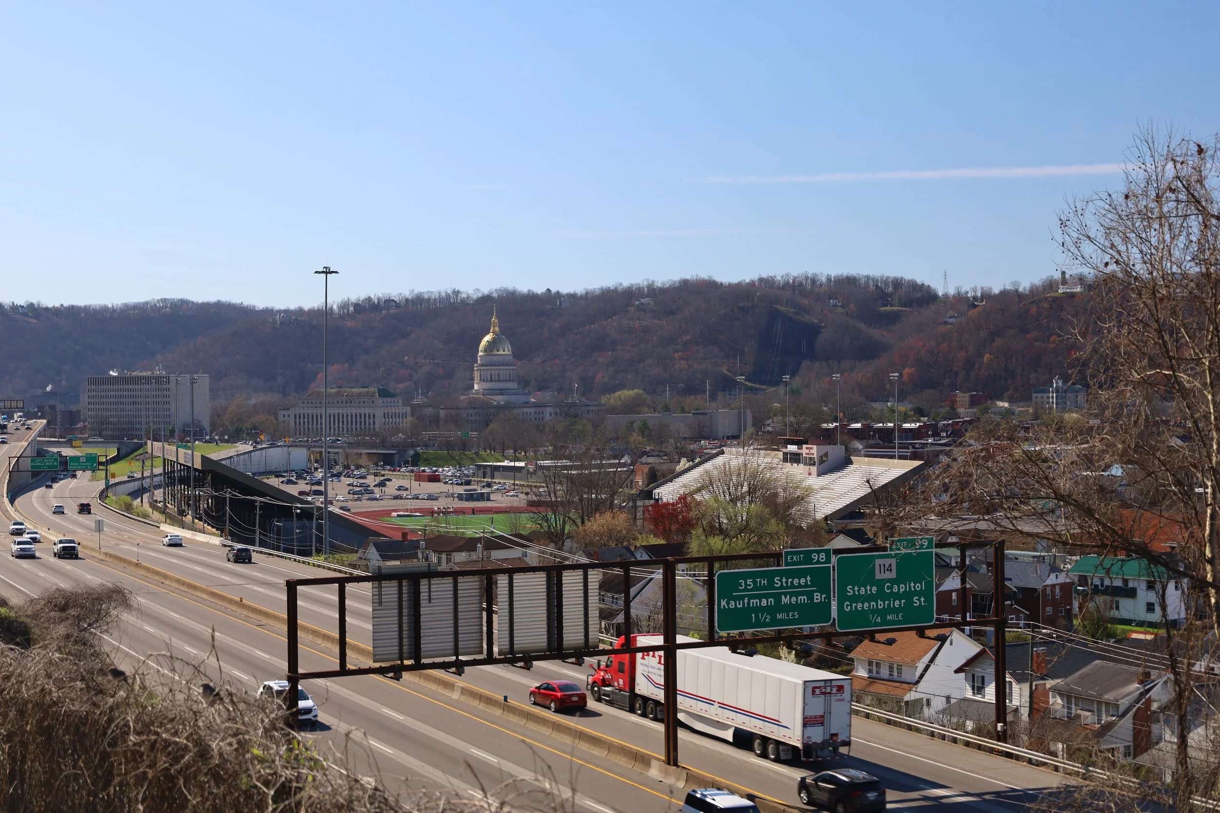 View of highway with cars, a traffic sign indicating the State Capitol building with its gold dome in the background, situated amidst hills.