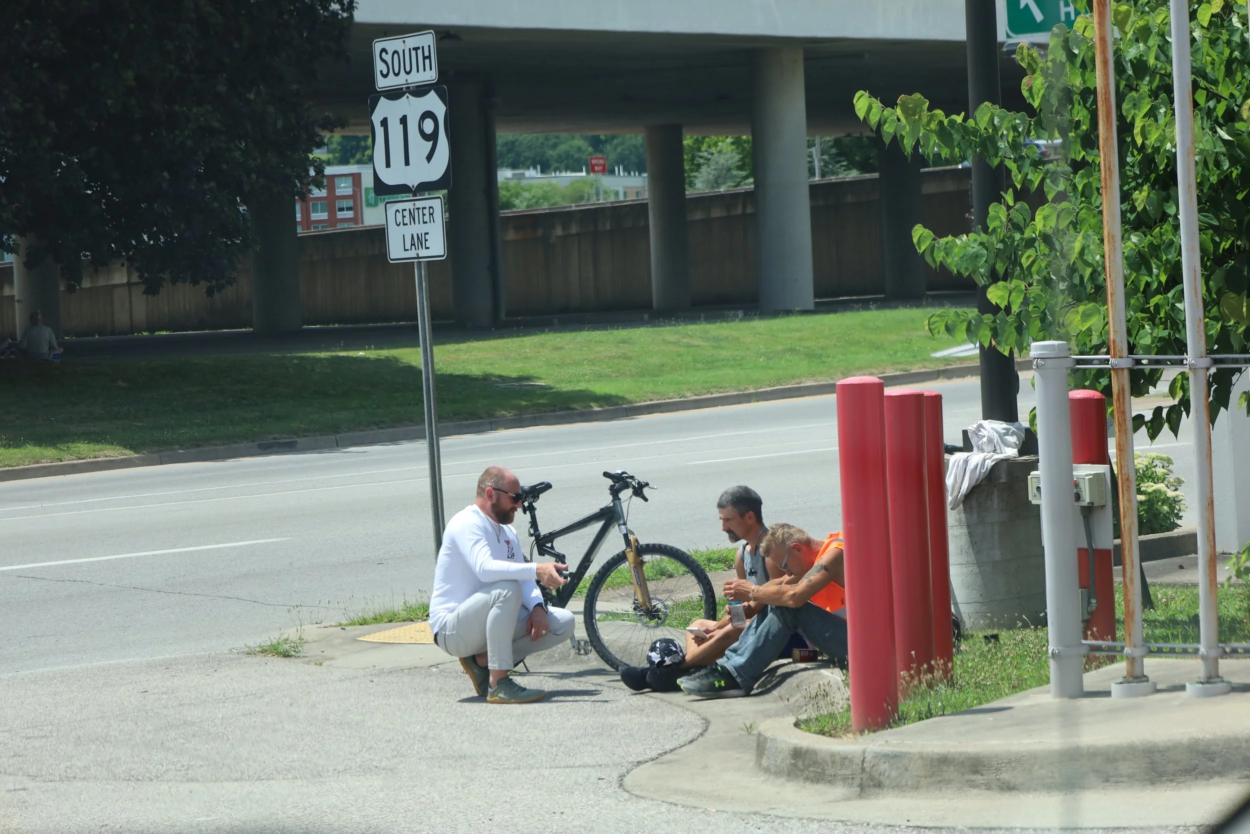 Three men sitting on the sidewalk near a bicycle and a sign for US Route 119 South.