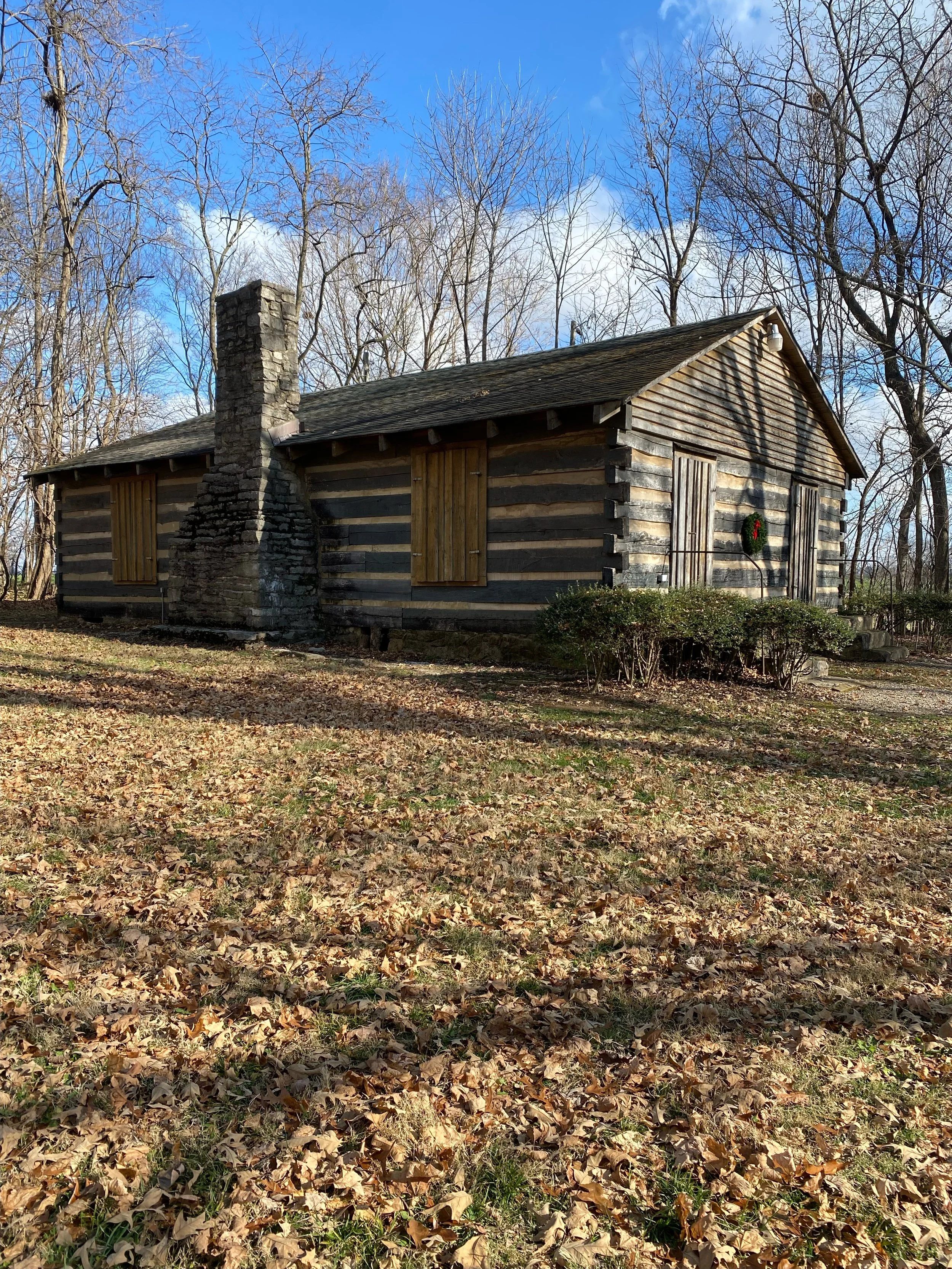 A rustic log cabin with shuttered windows and a stone chimney, surrounded by leafless trees and fallen leaves.