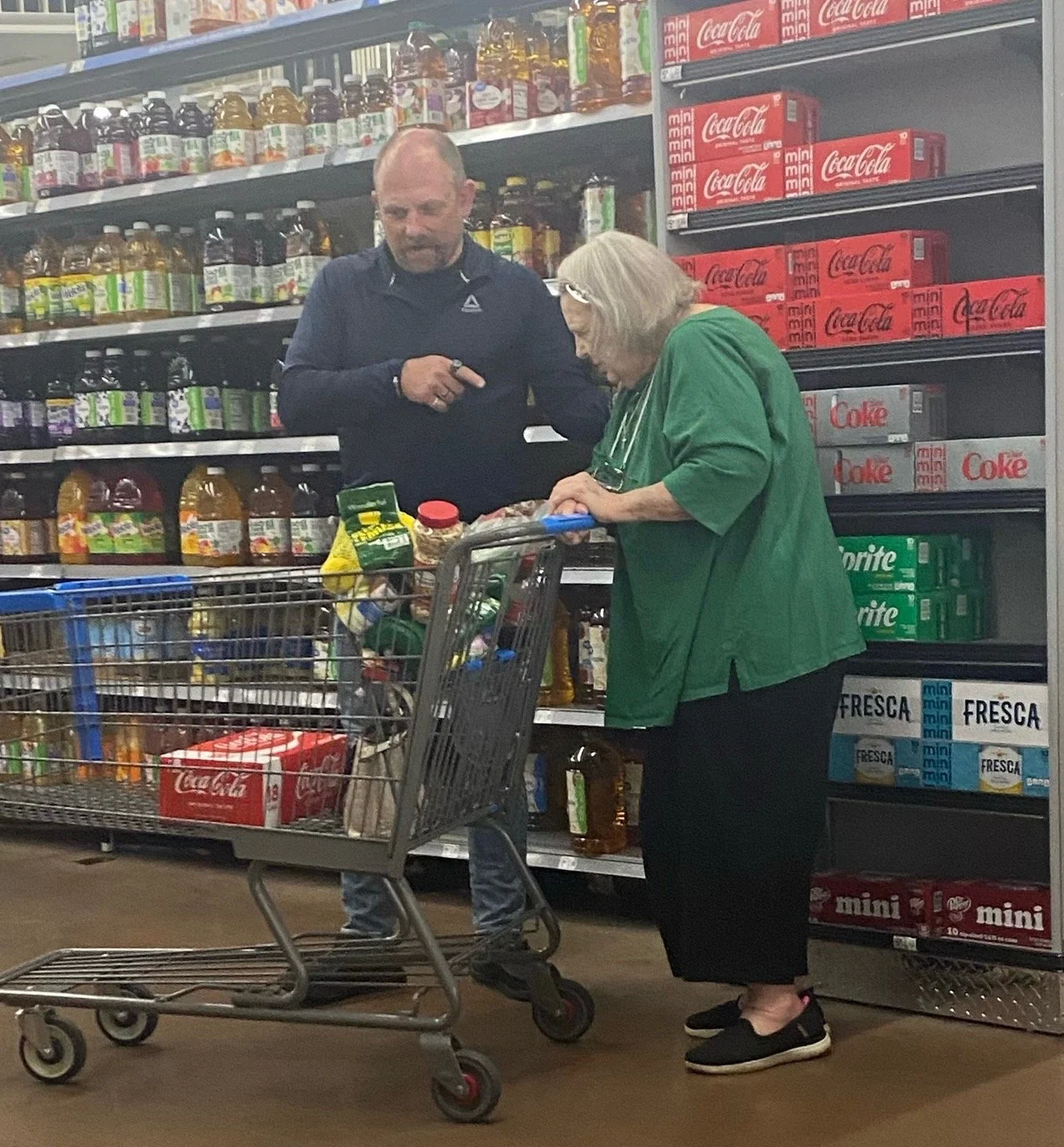 A man is praying for a woman shopping in a grocery store aisle, looking at products on the shelves. The woman is pushing a shopping cart filled with various items.