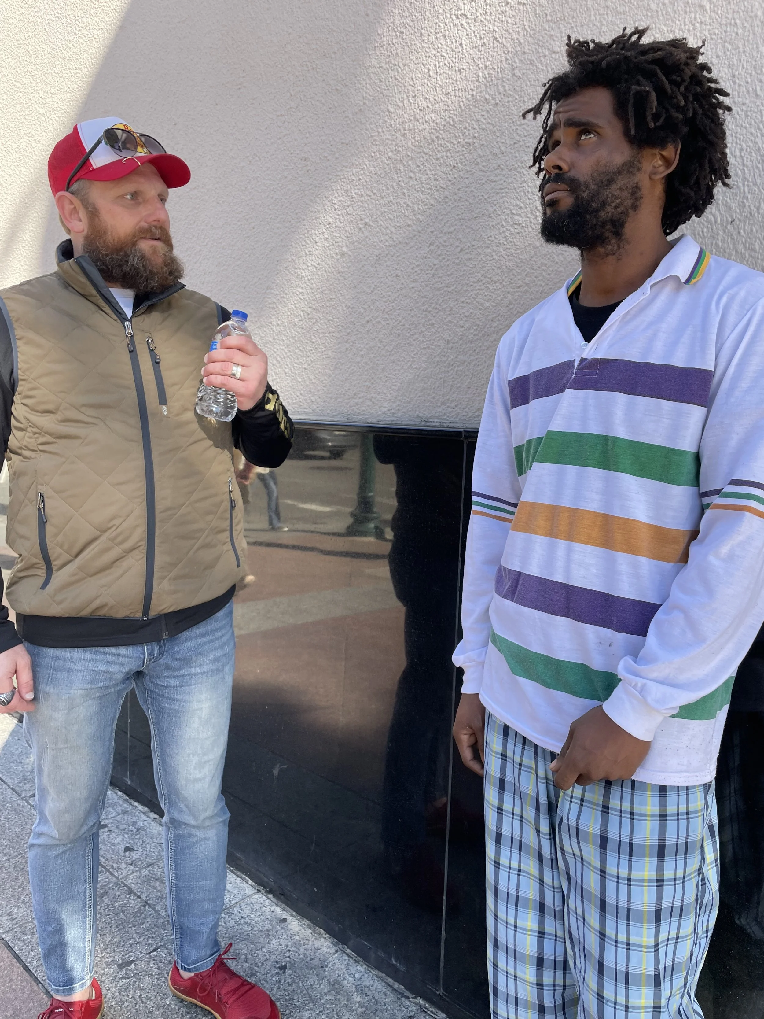 Two men standing outdoors, one holding a water bottle and the other with hands in pockets, engaged in conversation. The man on the left wears a red and white cap, sunglasses, a tan quilted vest, and jeans. The man on the right has dreadlocks, a beard