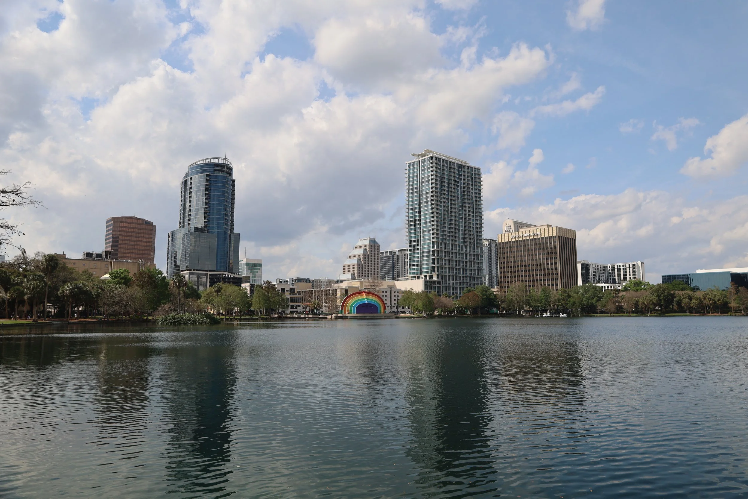 City skyline with modern high-rise buildings across a lake under a partly cloudy sky, with a rainbow mural on a building near the water.