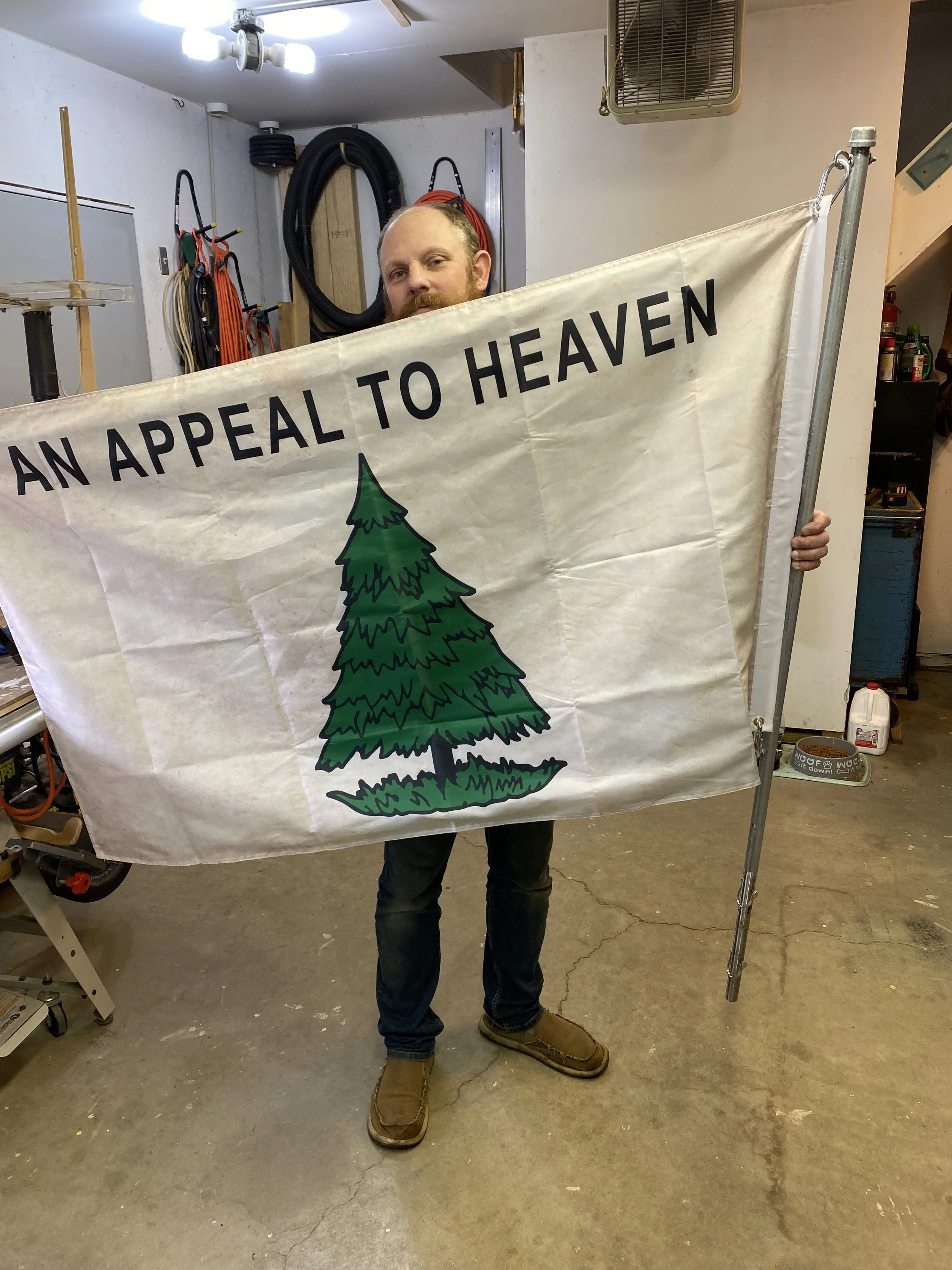 A man holding a banner with a green pine tree and the text 'An Appeal to Heaven' in a workshop.