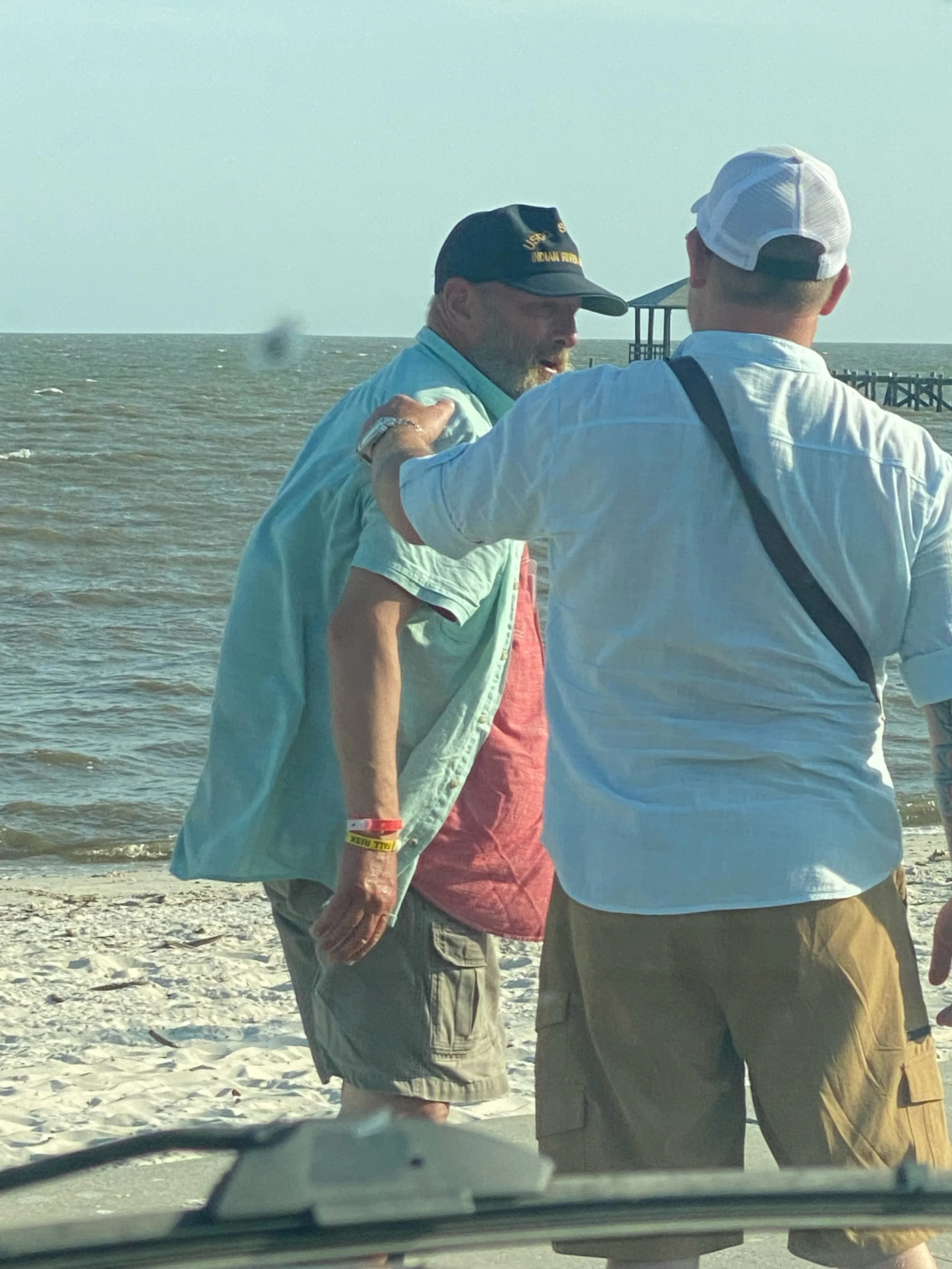 Two men stand near the beach, one with an army cap with yellow text, the other wearing a white cap, engaged in prayer, with a pier and water in the background.