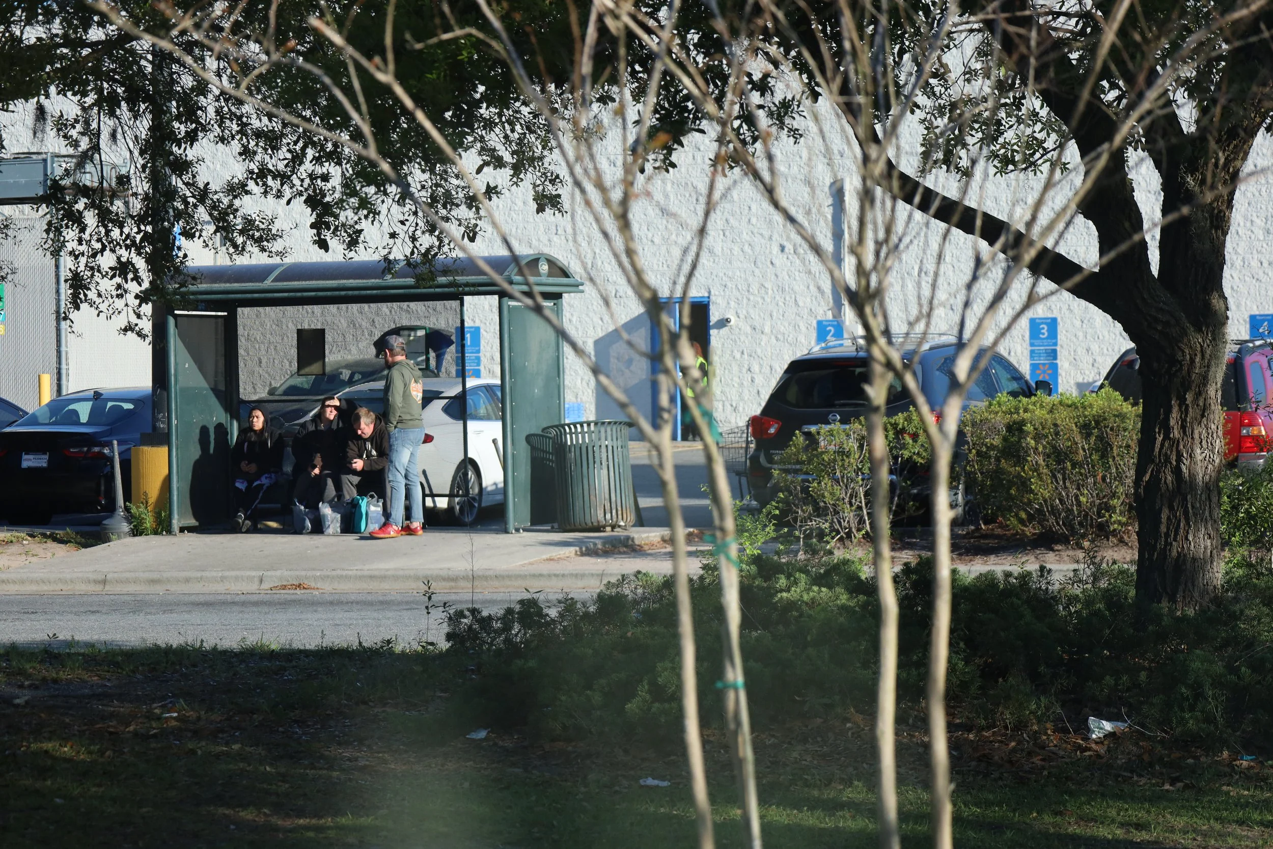 A group of people waiting and praying at a bus stop in a parking lot, with parked cars, trees, and a white building in the background.
