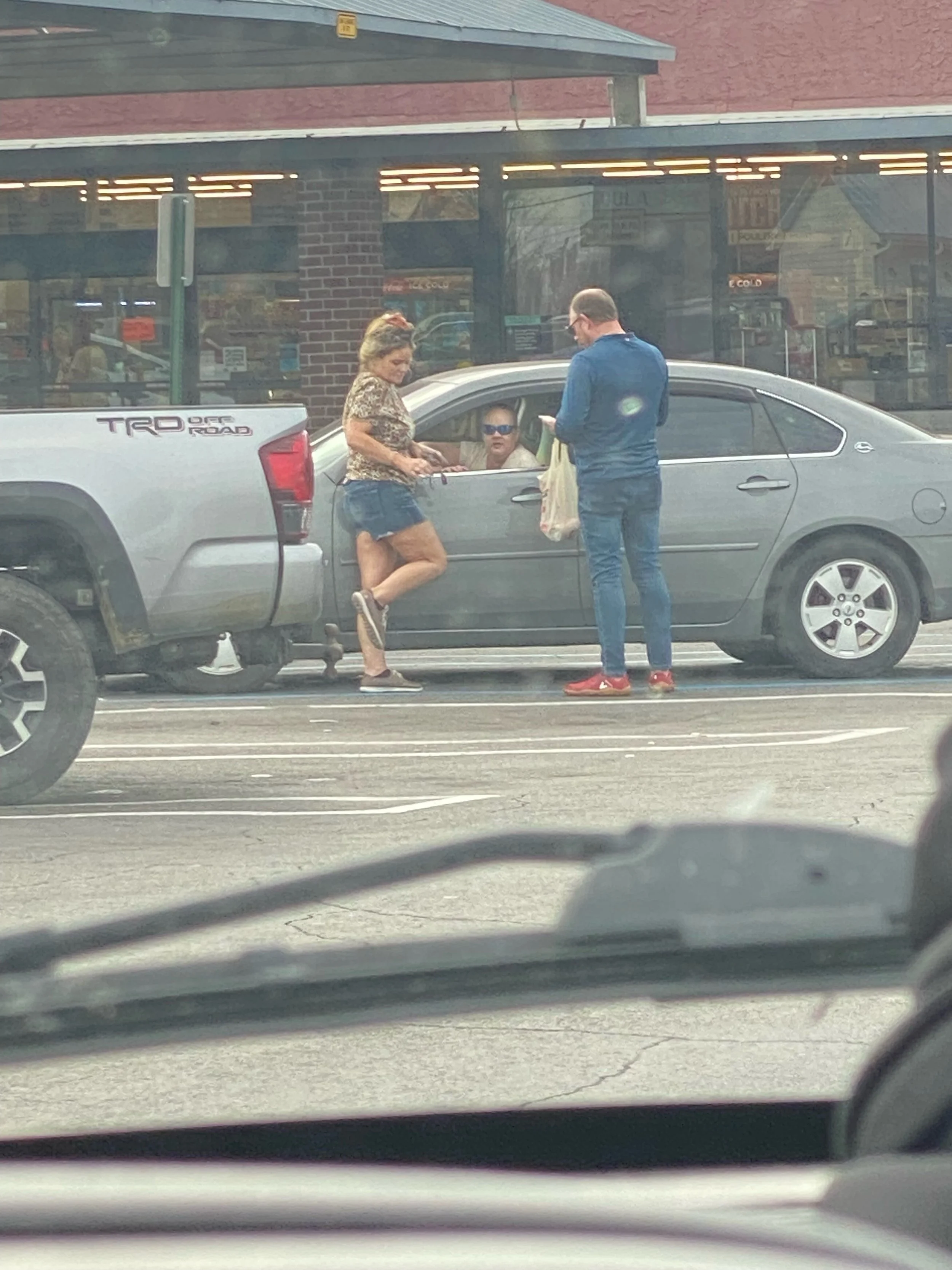A woman is speaking to a man standing outside holding a bag, while a woman inside the car looks on. The woman is moving her knee without pain after prayer. The scene is set in a parking lot outside a store.