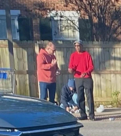 Two men standing and talking and praying outside near a wooden fence, with a person sitting on the ground in front of them.