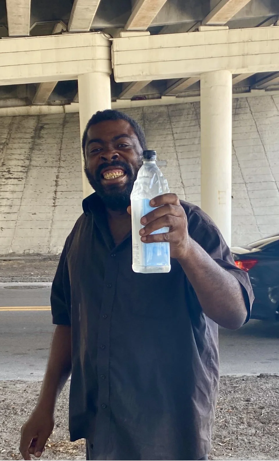 A man smiling and showing his teeth while holding a plastic water bottle, standing outdoors under an overpass.