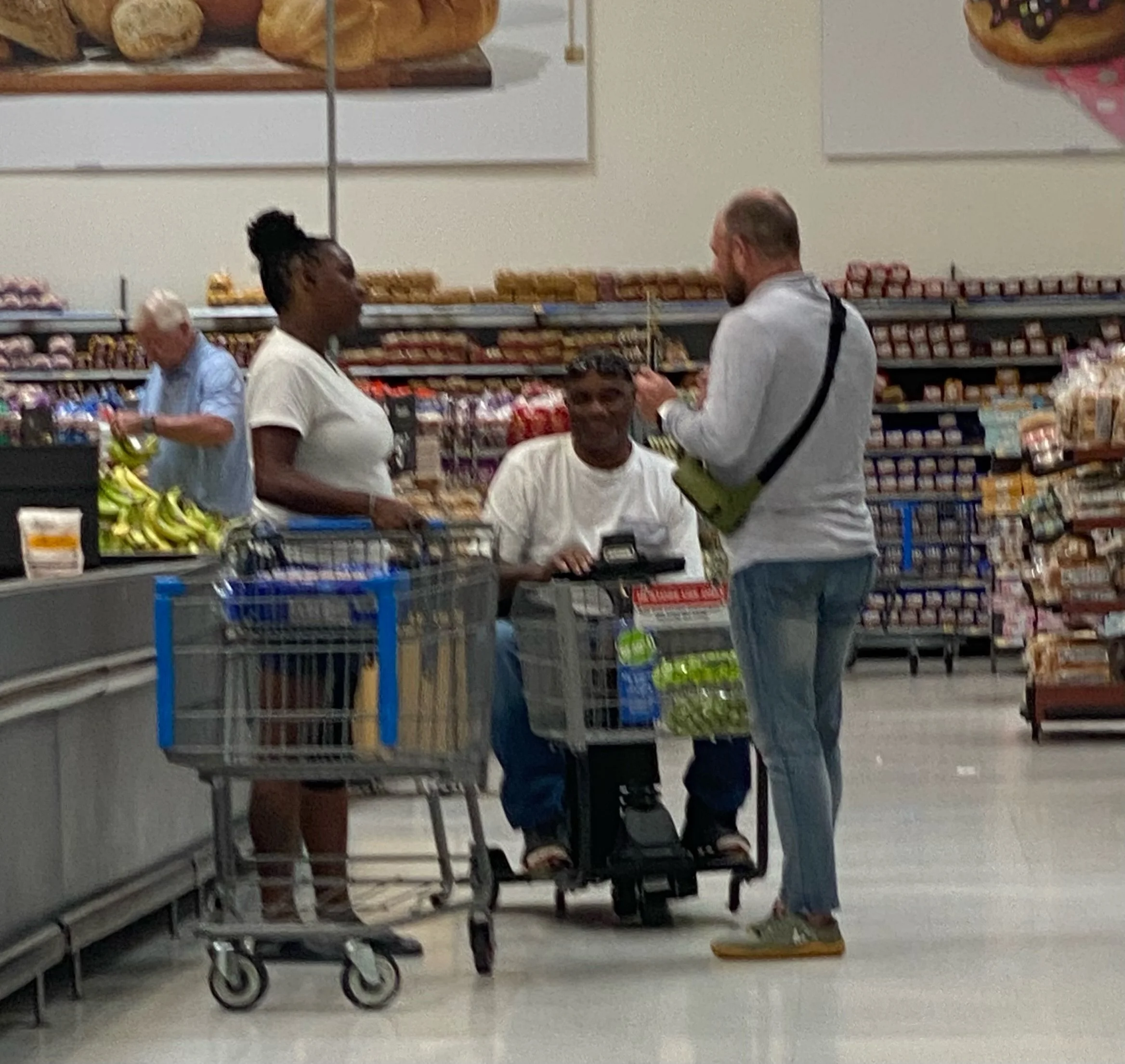 Three people at a grocery store. One person is sitting in a motorized cart, and two others are standing beside and in front of him. A woman is in line, the man is praying and an older woman is shopping in the background.
