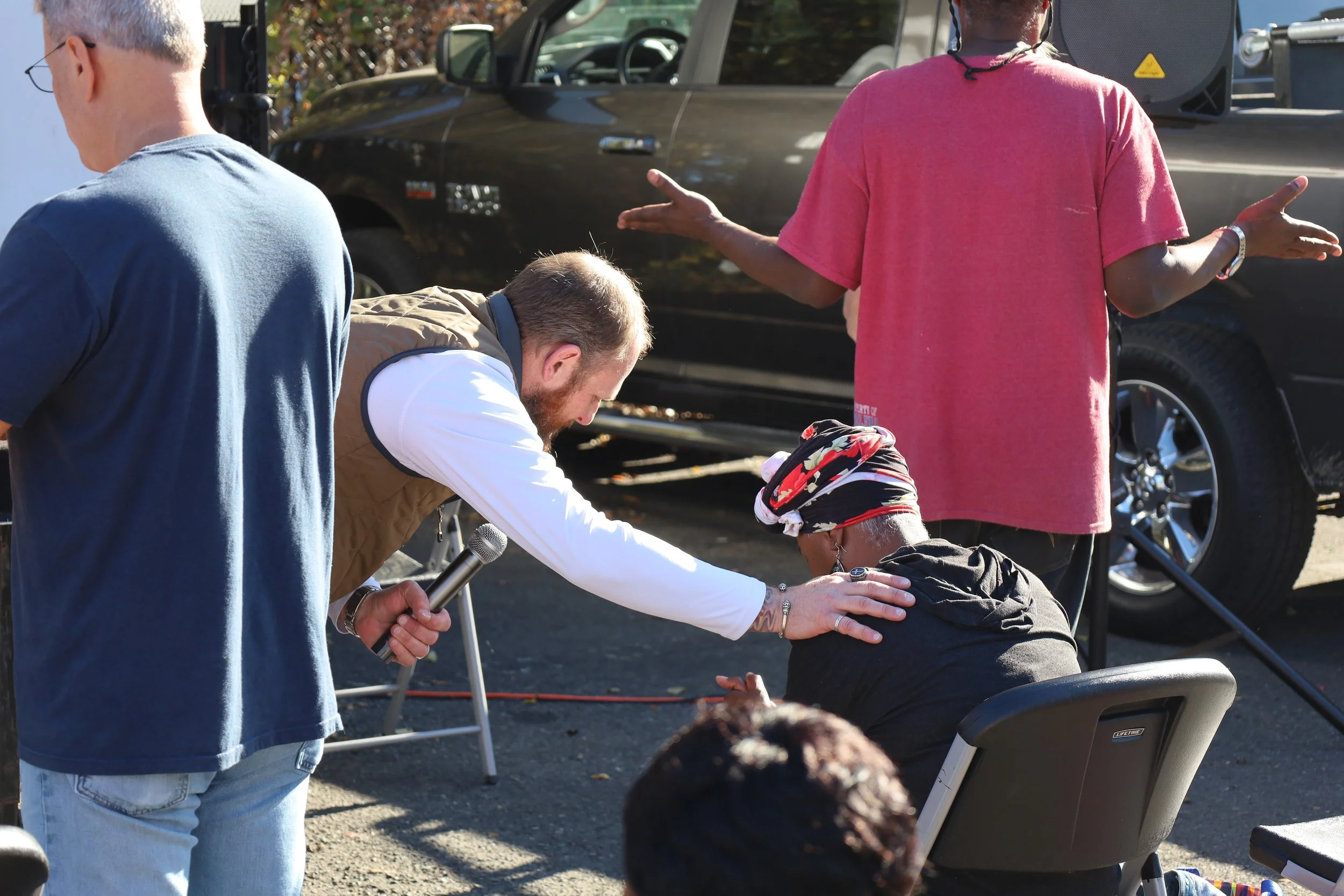 A woman sitting in a chair being being prayed for by a man, while interacting with a person in a pink shirt with arms outstretched, outdoors next to a black vehicle.