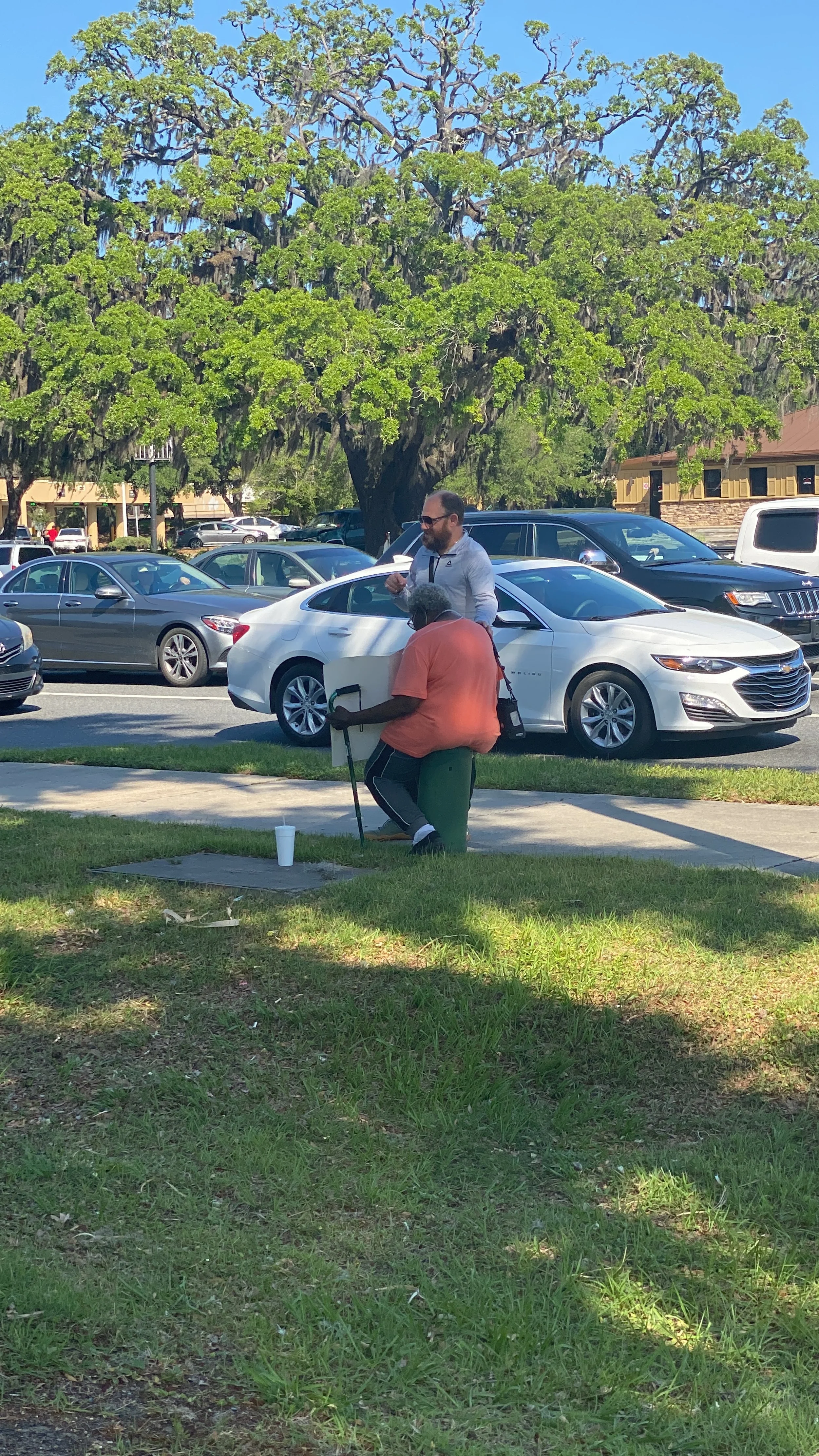 A man and a woman are outside praying under a large leafy tree, with parking lot and cars in the background.