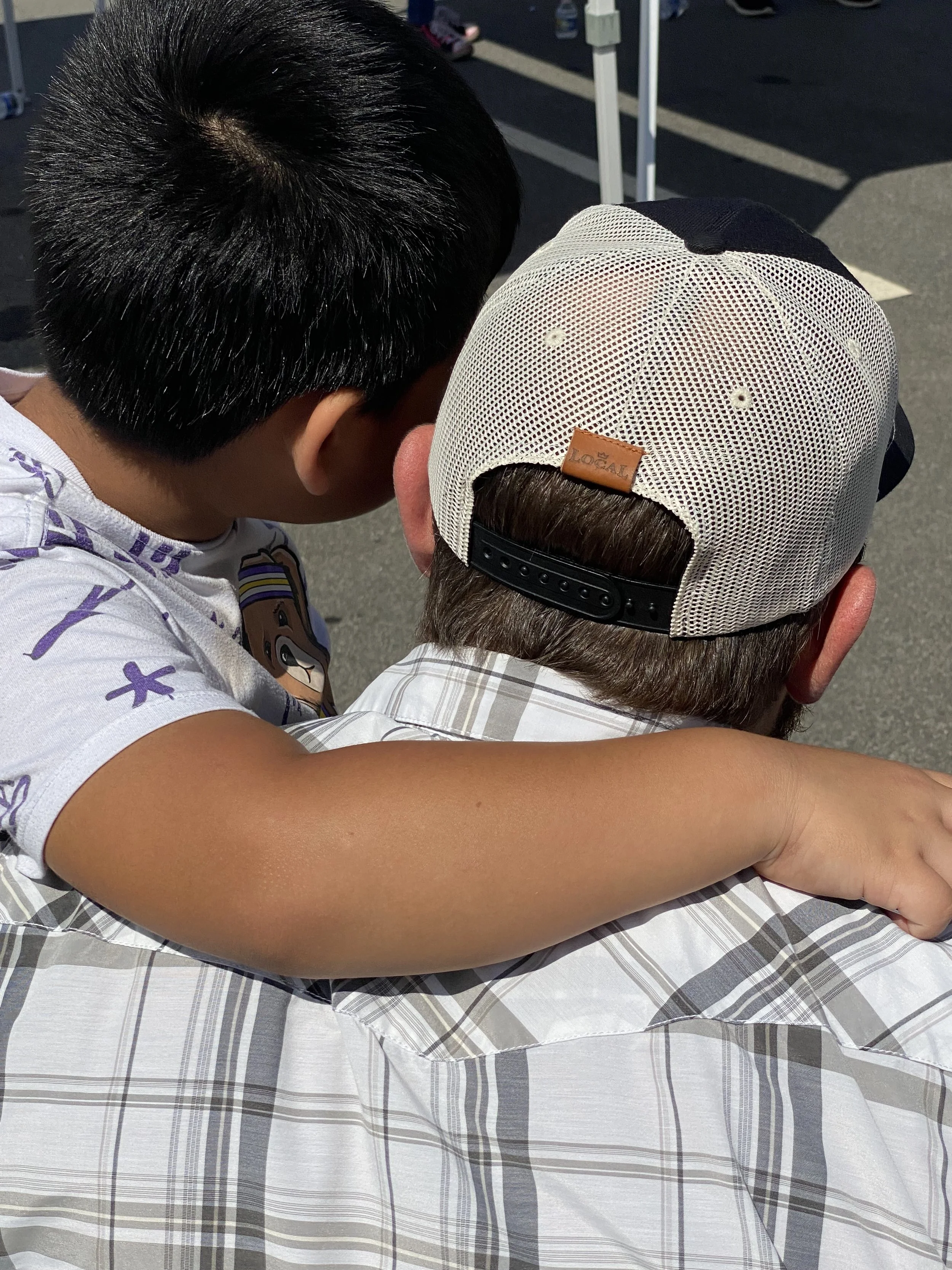 A young boy hugging an older man, with his arm around the man's neck, in an outdoor parking lot. The boy has short black hair and is wearing a white t-shirt with cartoon characters. The man is wearing a plaid shirt and a white mesh cap.