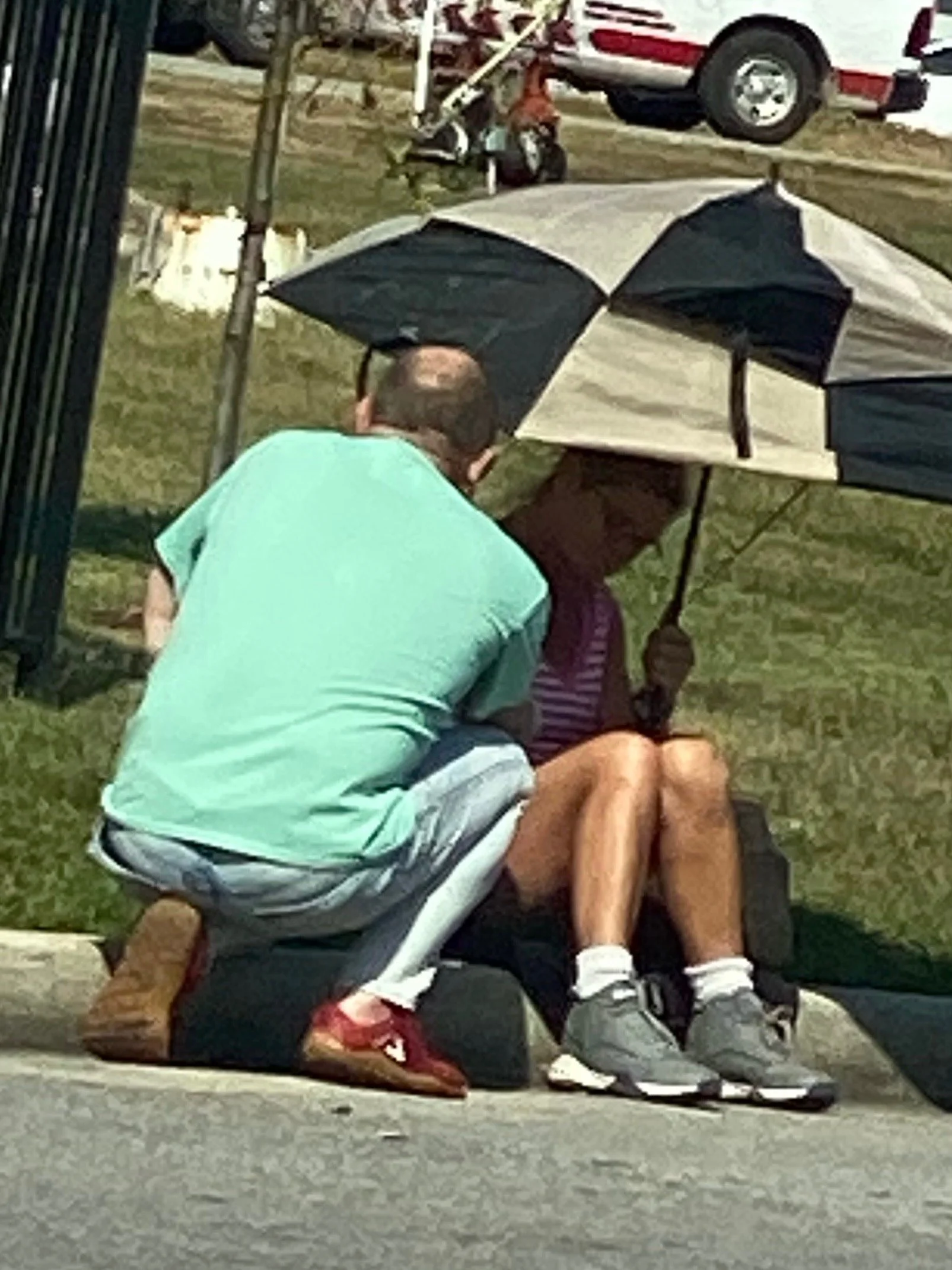 A man in a mint green shirt and gray shorts kneels next to a woman sitting on the sidewalk, holding an umbrella. The woman is wearing a striped shirt, shorts, and sneakers. They are engaged in conversation beneath the umbrella, with a parked car and 