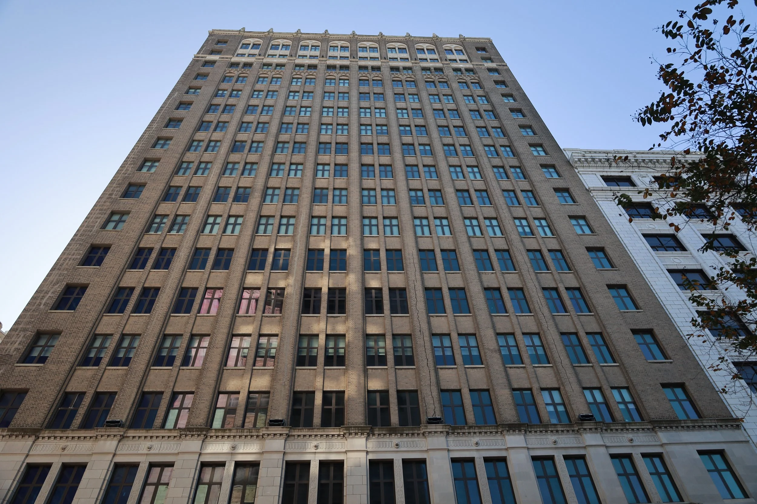 A tall multi-story building with a brick facade and numerous windows, viewed from the ground looking upward. Part of a neighboring building and tree branches are visible on the right side.
