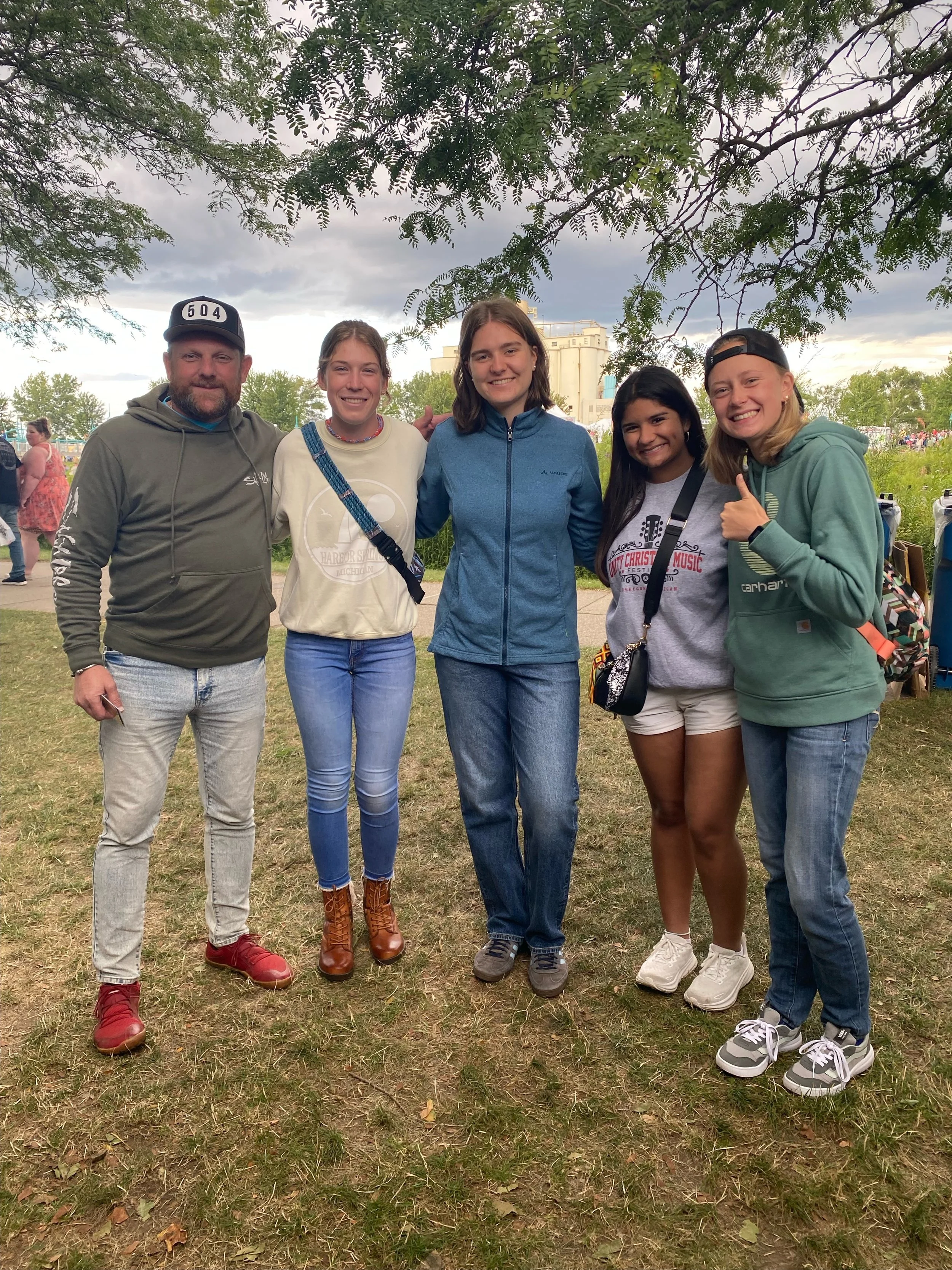 Group of five people standing outdoors on grass, smiling at the camera. Overcast sky with dark clouds in the background, trees and a building visible behind them.