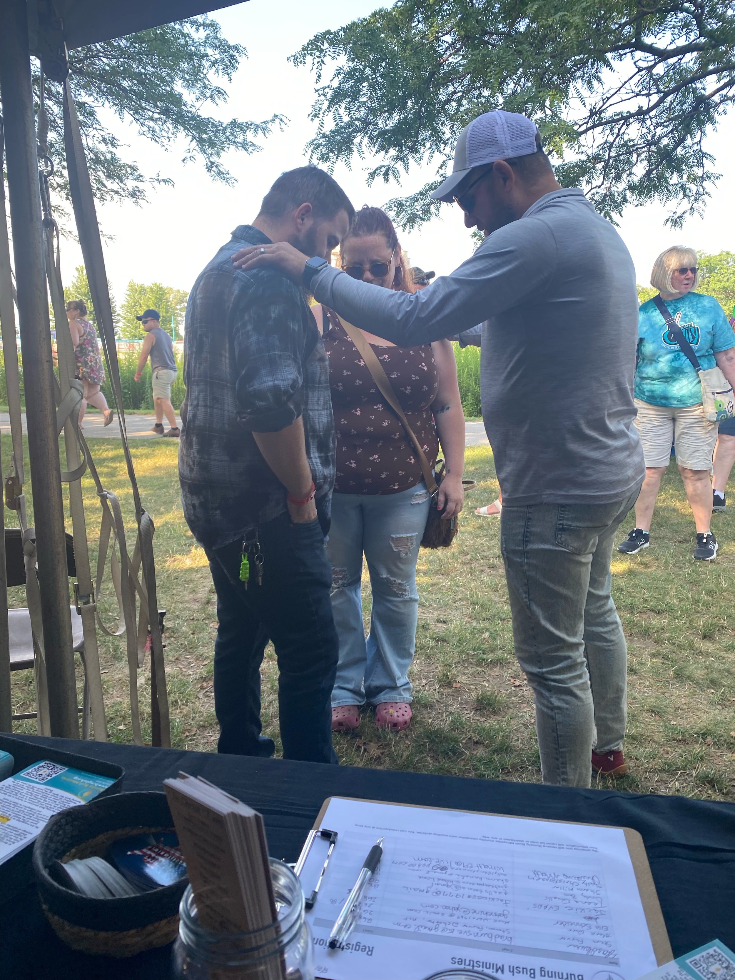 Three people are standing outdoors under a tree, with their heads bowed and hands on each other's shoulders, in a prayer. In the background, there are other people walking and standing on the grassy area.