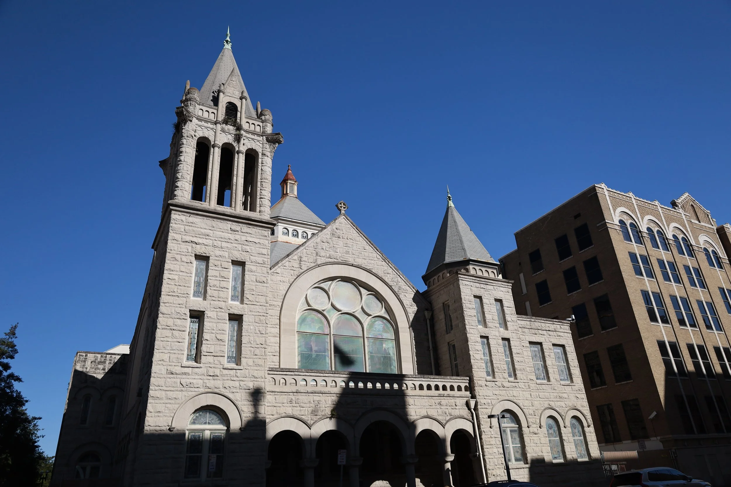 A historic church with gothic architecture, stone walls, stained glass windows, and tall towers topped with spires, set against a bright blue sky.